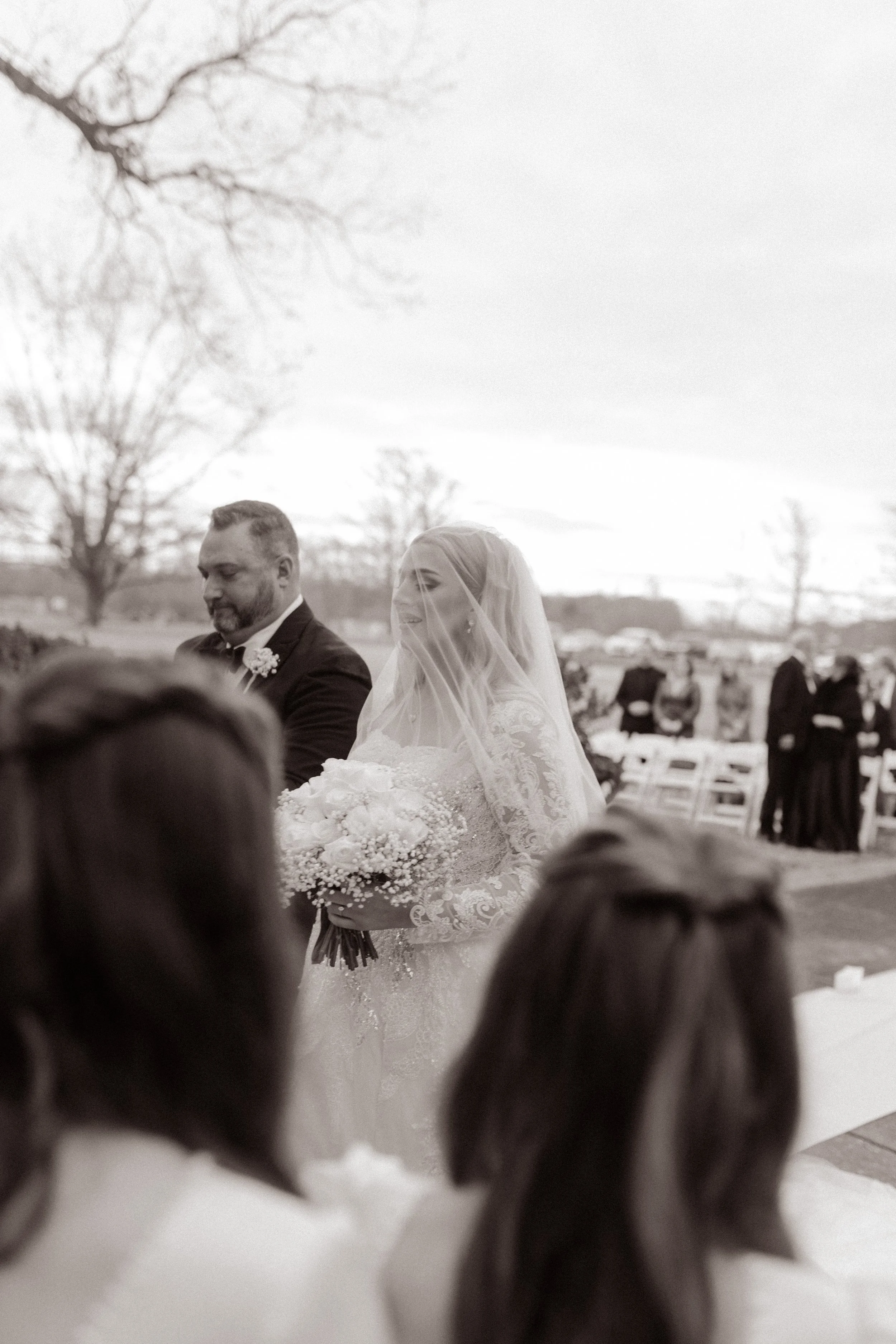 Bride walks down the aisle on her father’s arm, bouquet in hand and veil over her face, with guests standing in the background.