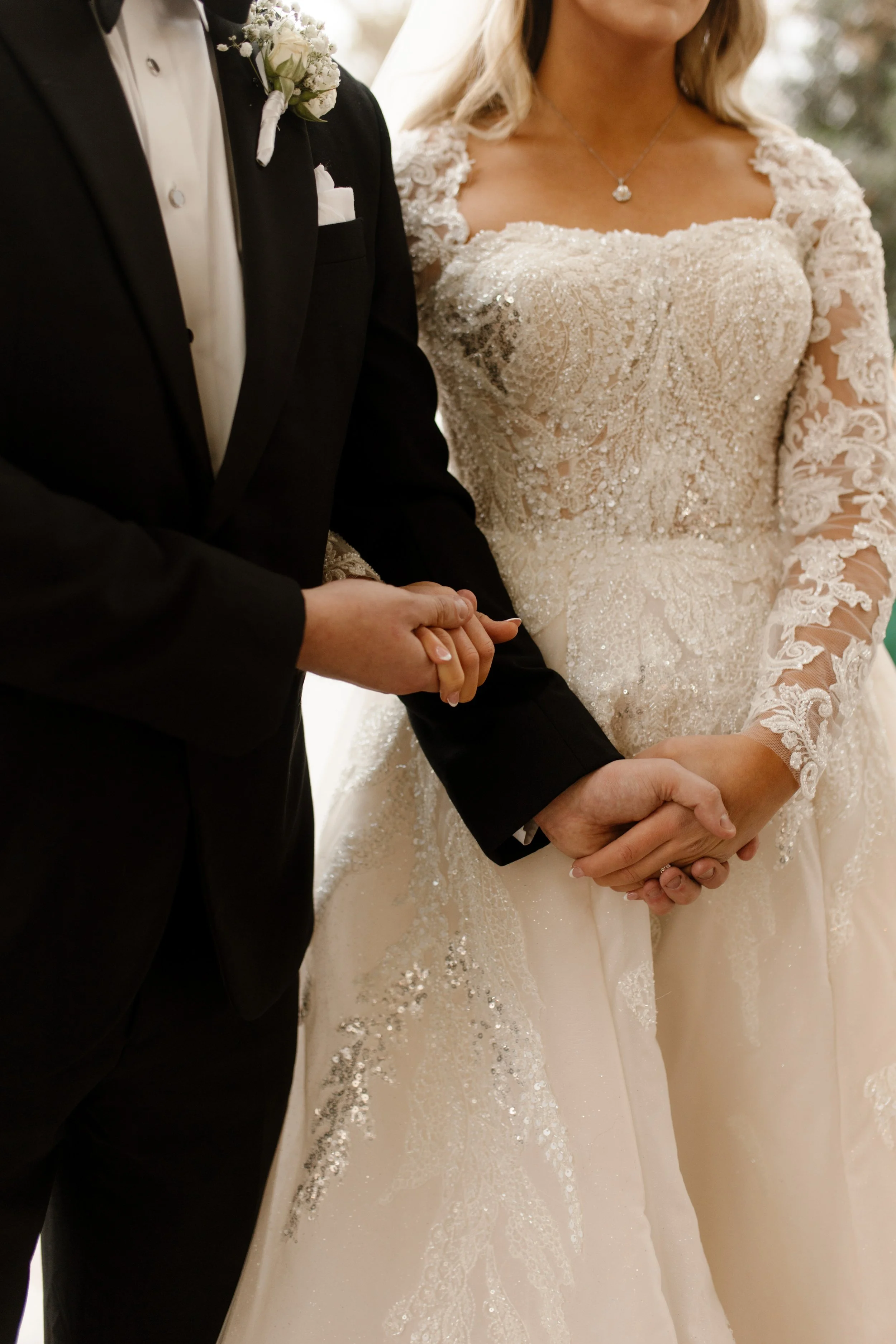 Detail shot of the bride and groom’s intertwined hands during the ceremony, her beaded lace sleeve and his tux jacket meeting in a quiet, powerful moment.
