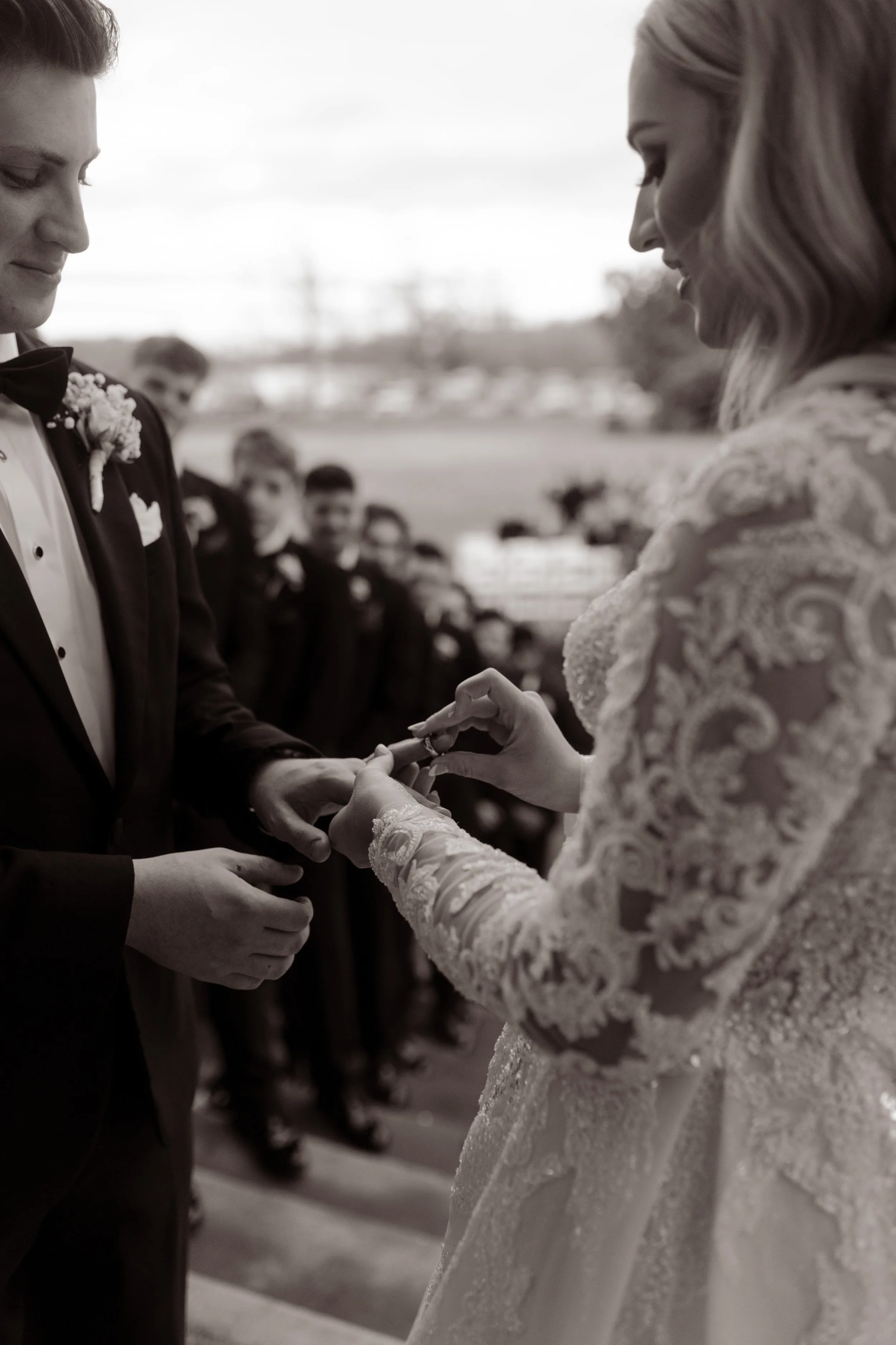 During the ceremony, the bride gently places a ring on her groom’s hand, framed by rows of guests and soft outdoor light at this Arkansas wedding.