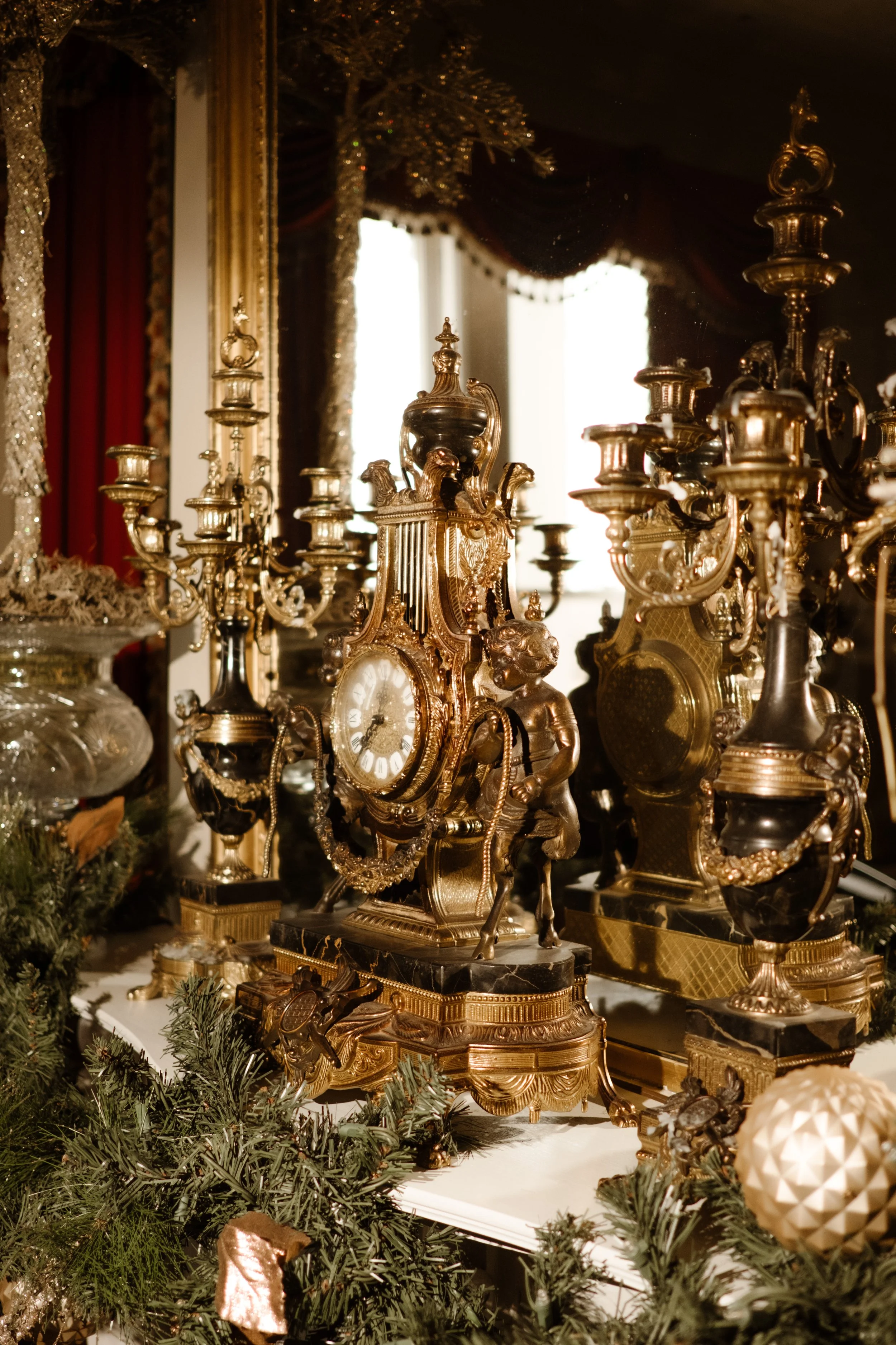 Close-up of ornate antique decor—gilded clock and candle holders resting on a mantle draped in evergreens and gold ornaments.