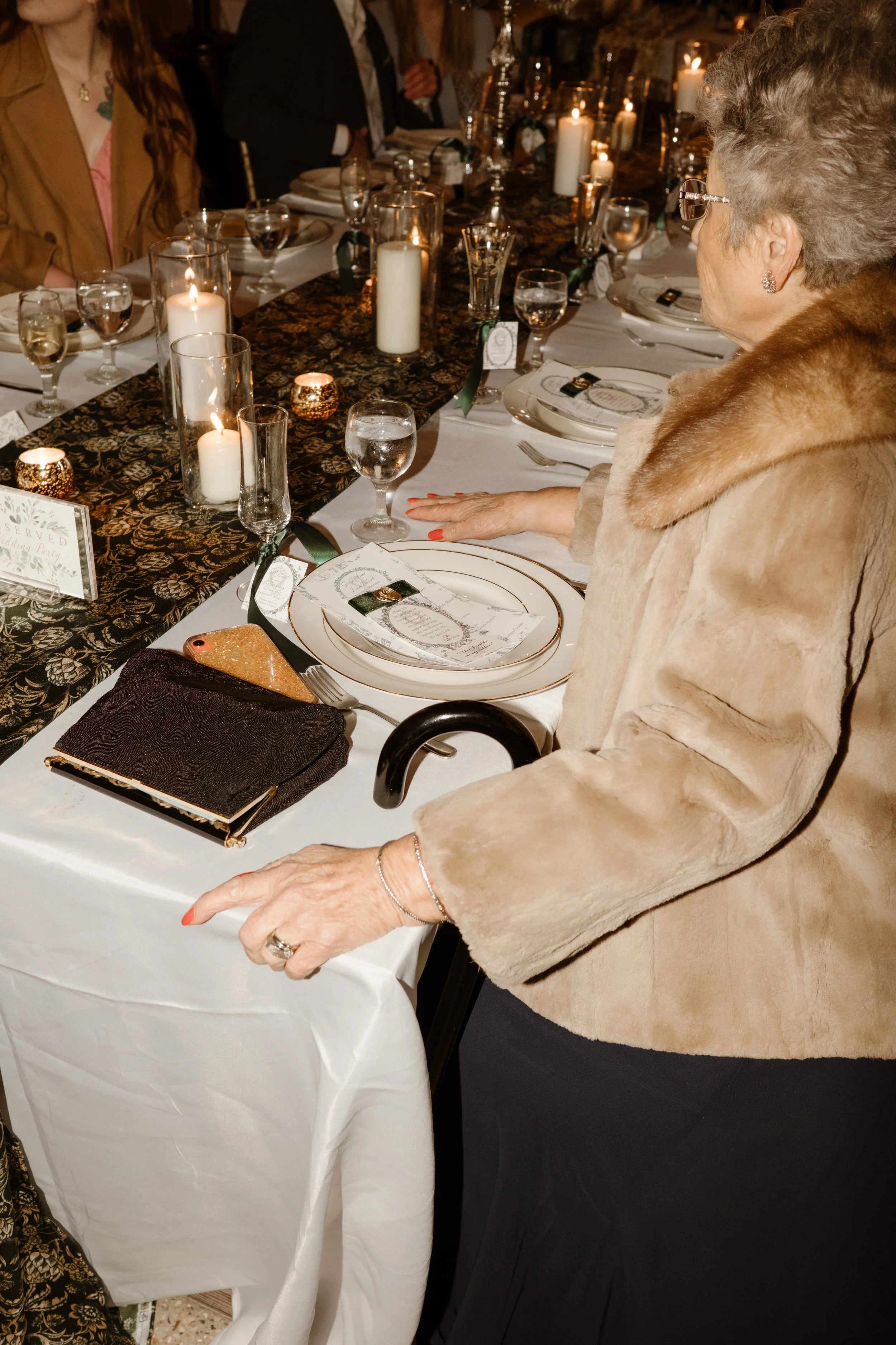 A guest in a fur-trimmed coat sits at a candlelit table, hands gently resting near a vintage clutch and custom place setting.
