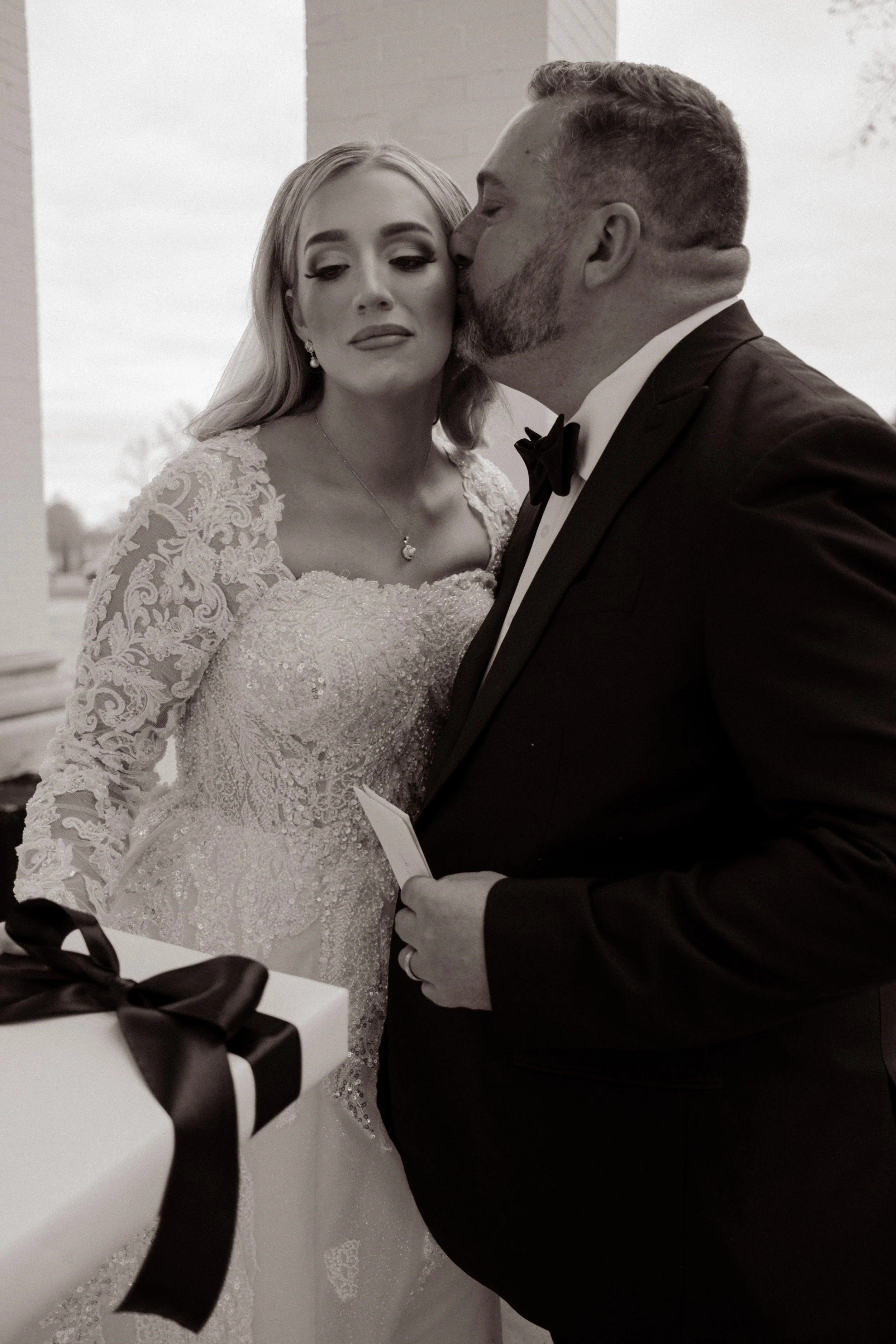Bride in a lace gown receives a tender kiss on the cheek from her father moments before the ceremony, holding back tears with a gift in hand.