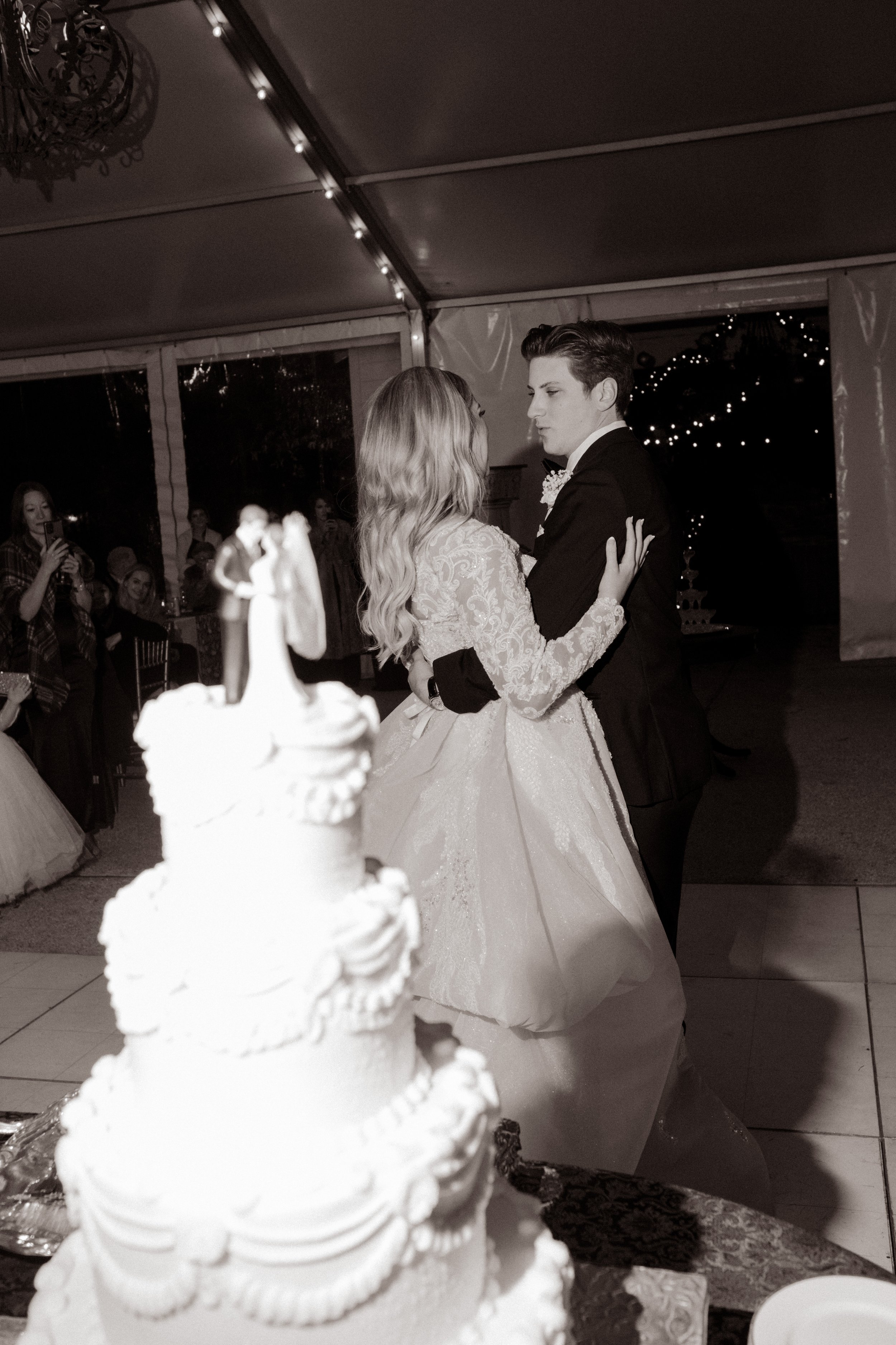 Bride and groom share their first dance under twinkle lights at their Arkansas wedding, framed by a towering vintage-style wedding cake in the foreground.