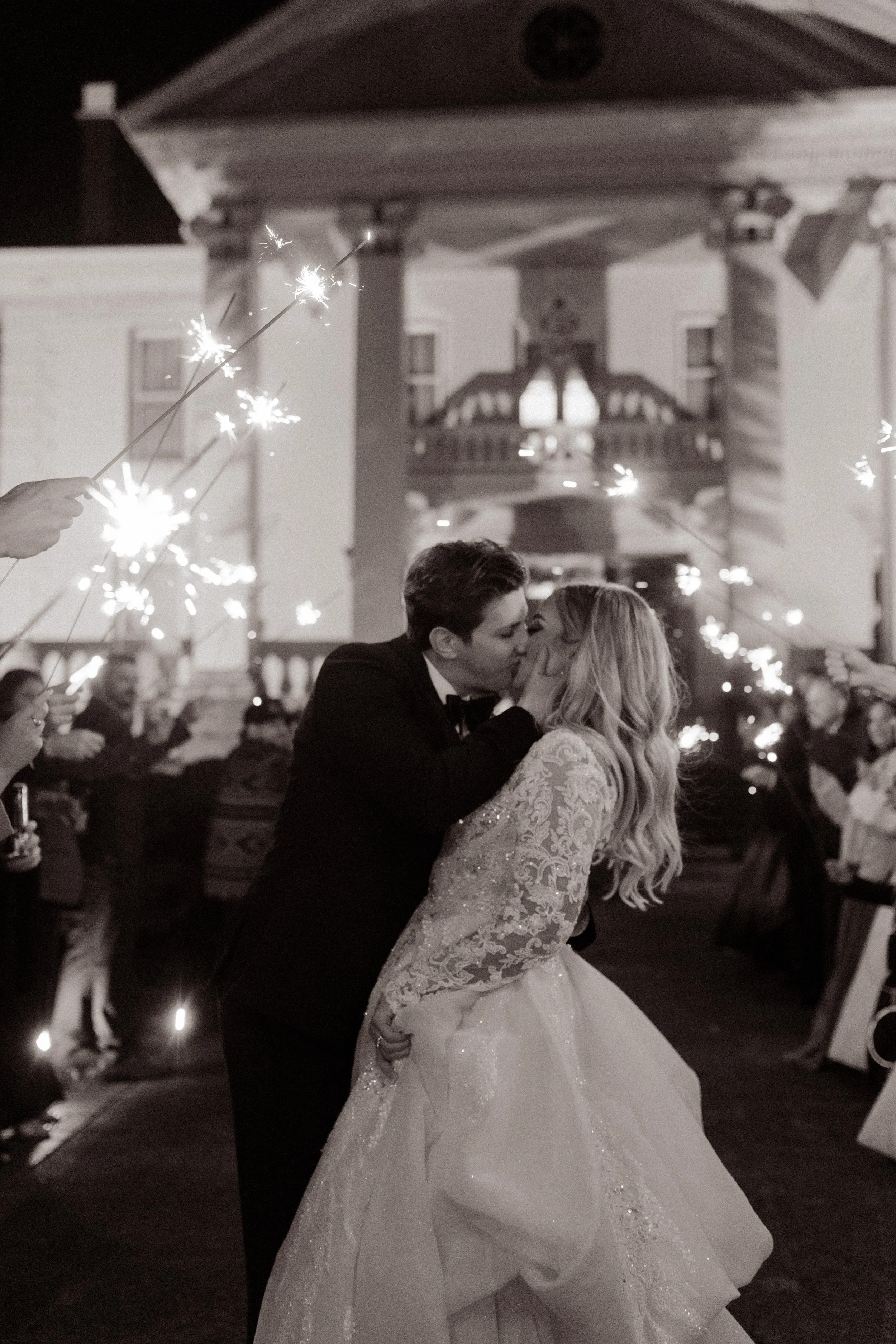Newlyweds share a dramatic kiss under a tunnel of sparklers as guests cheer outside a grand estate during their Arkansas wedding celebration.
