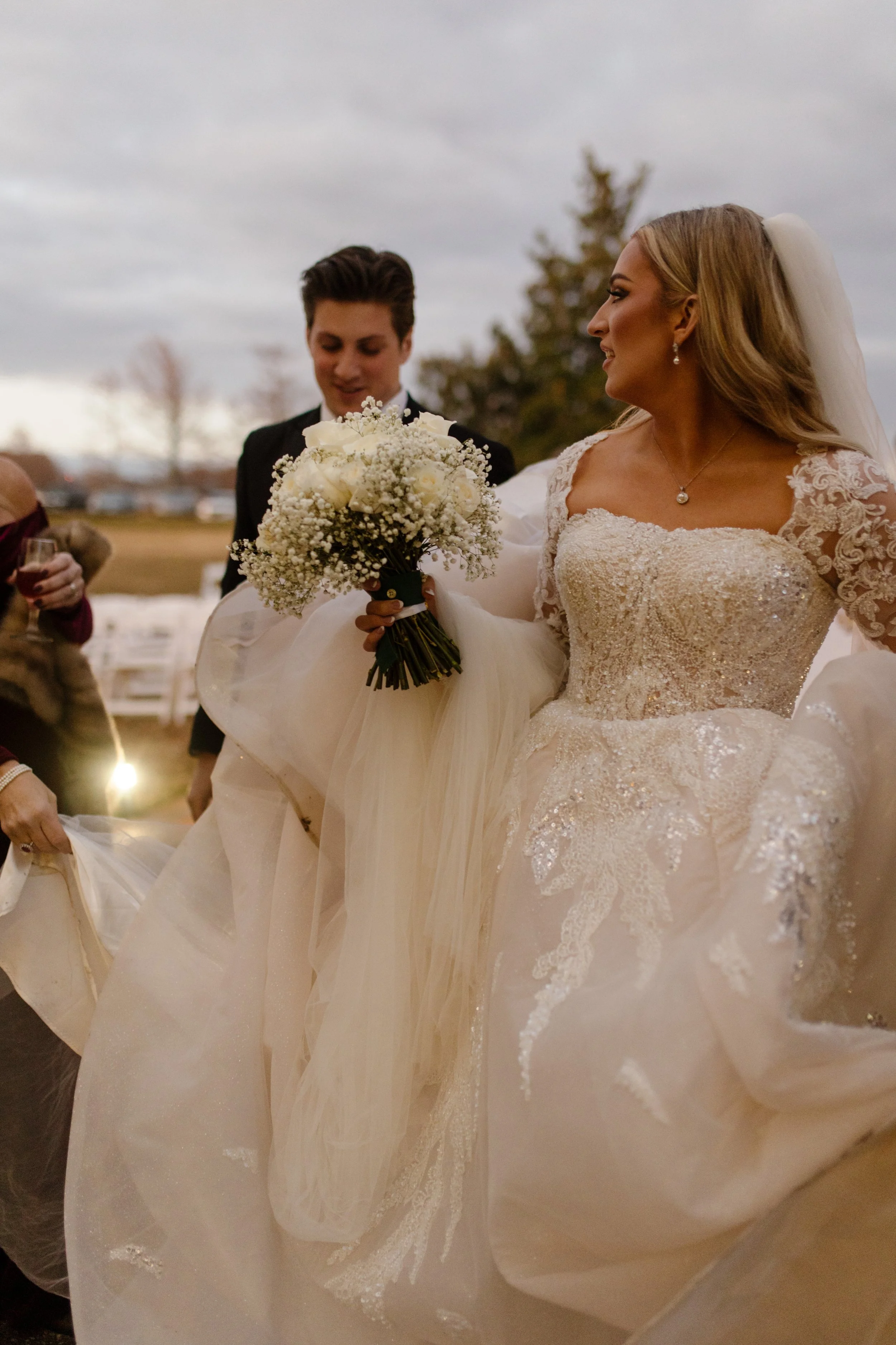 Bride clutching baby’s breath bouquet walks hand in hand with her groom, gown gathered in one hand as the evening sky sets a moody tone.