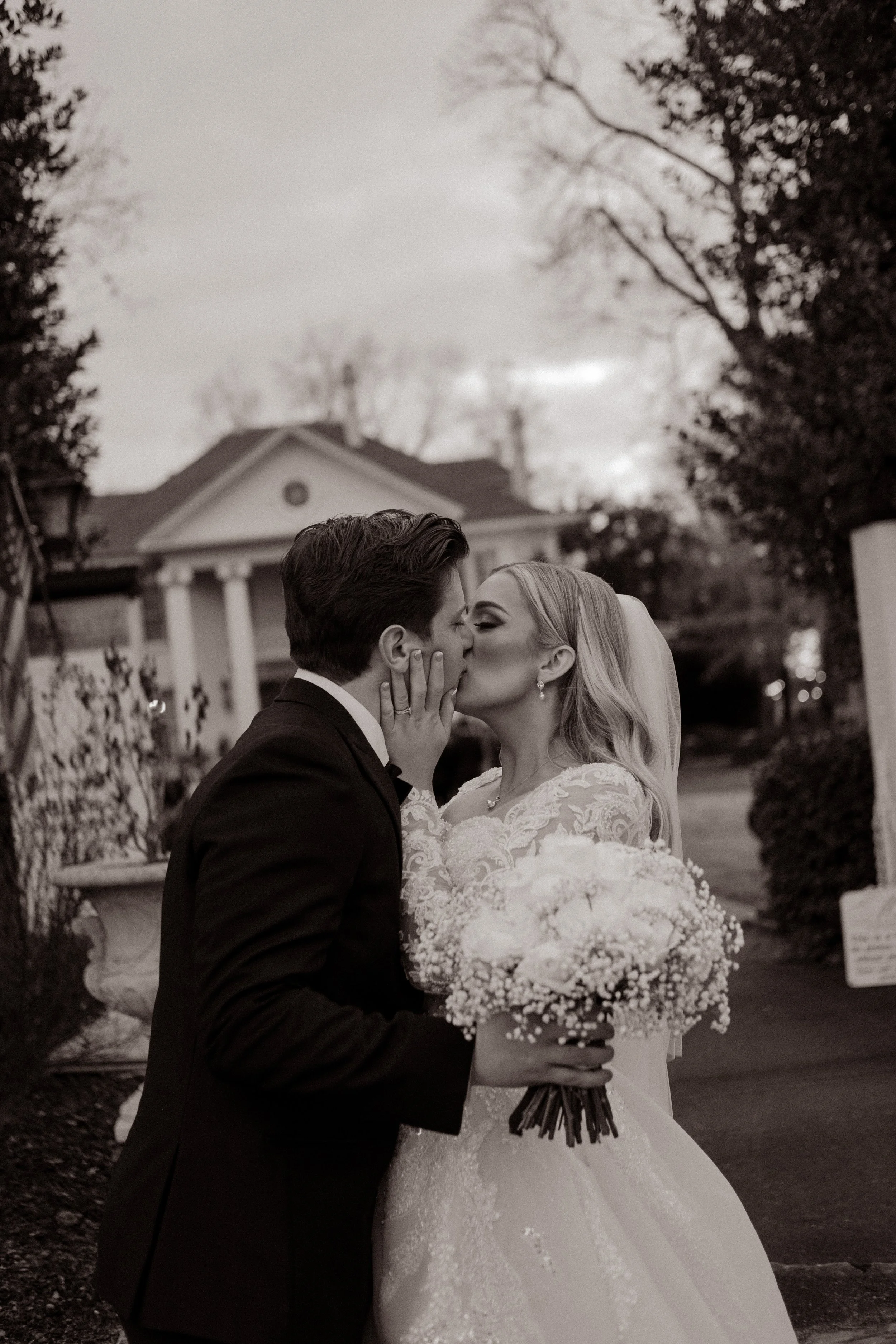 Bride and groom pause for a quiet kiss outside the venue, her hand on his cheek, bouquet in hand, framed by winter trees and soft dusk light.