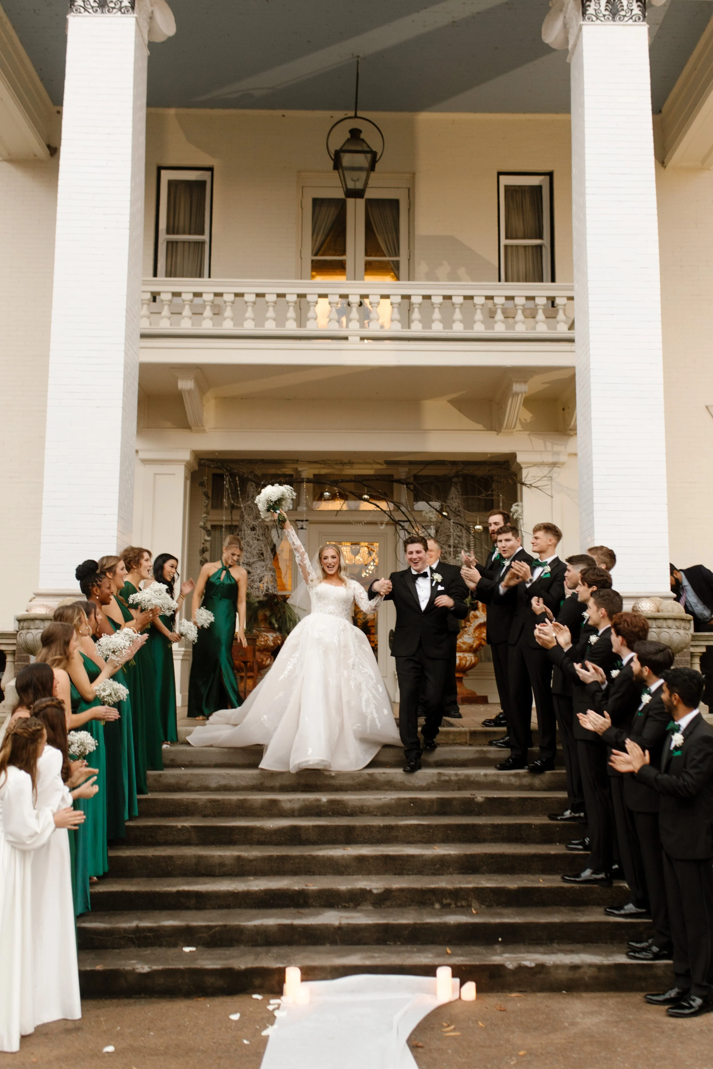 Bride and groom walk triumphantly down the steps of a white-columned estate, surrounded by their wedding party cheering them on during their Arkansas wedding.
