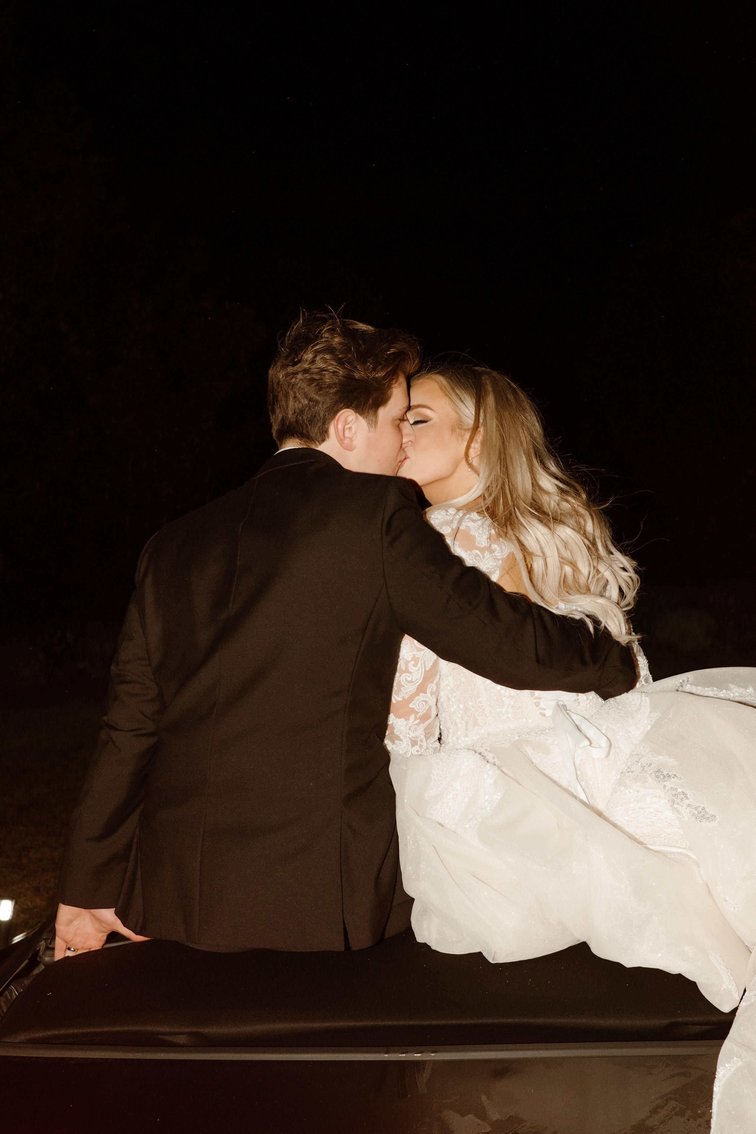 Couple sneaks one last kiss while perched on the back of their getaway car, dress flowing around them in the night air.