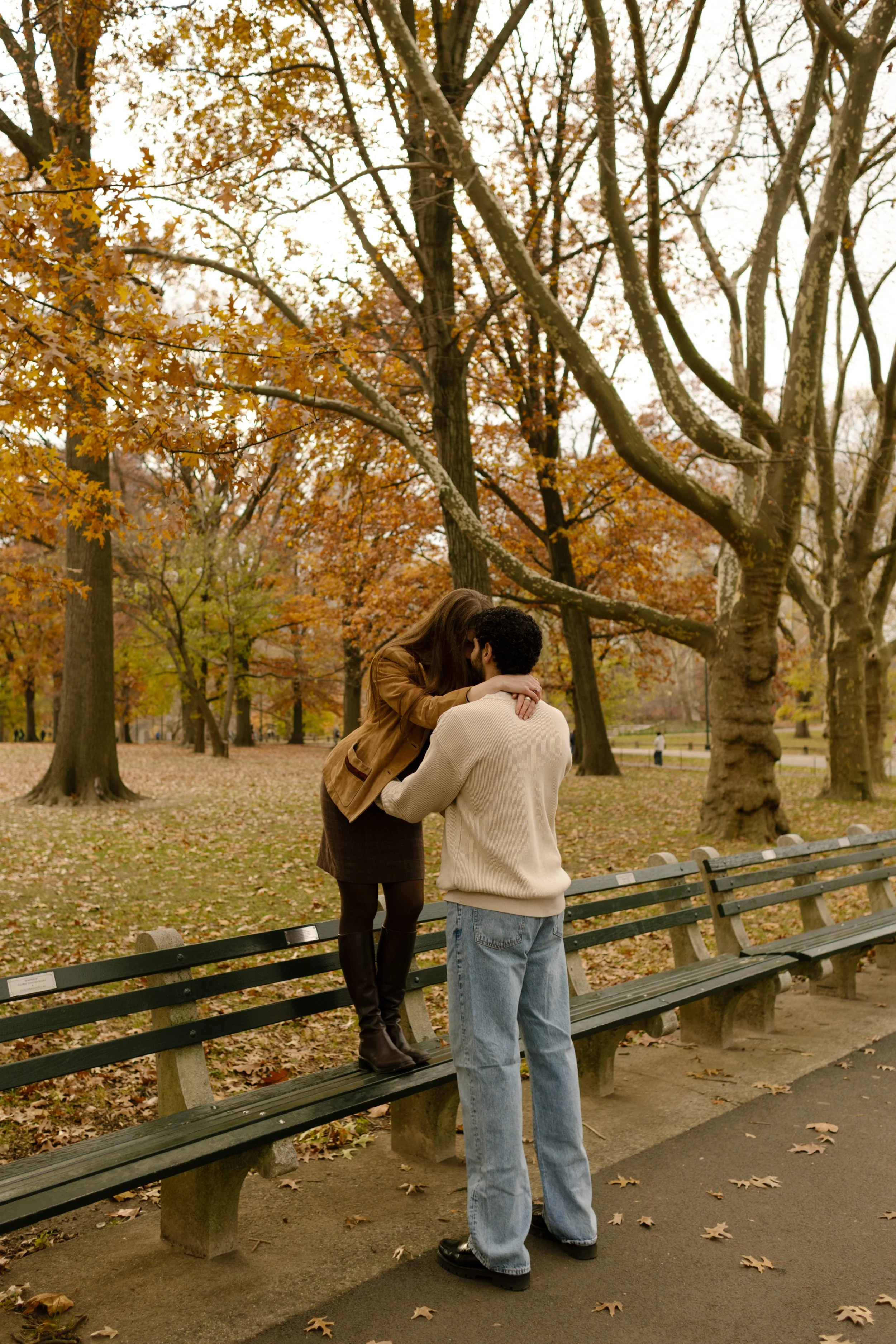 Woman standing on a bench kissing her partner in a cozy fall scene during their Central Park engagement shoot.