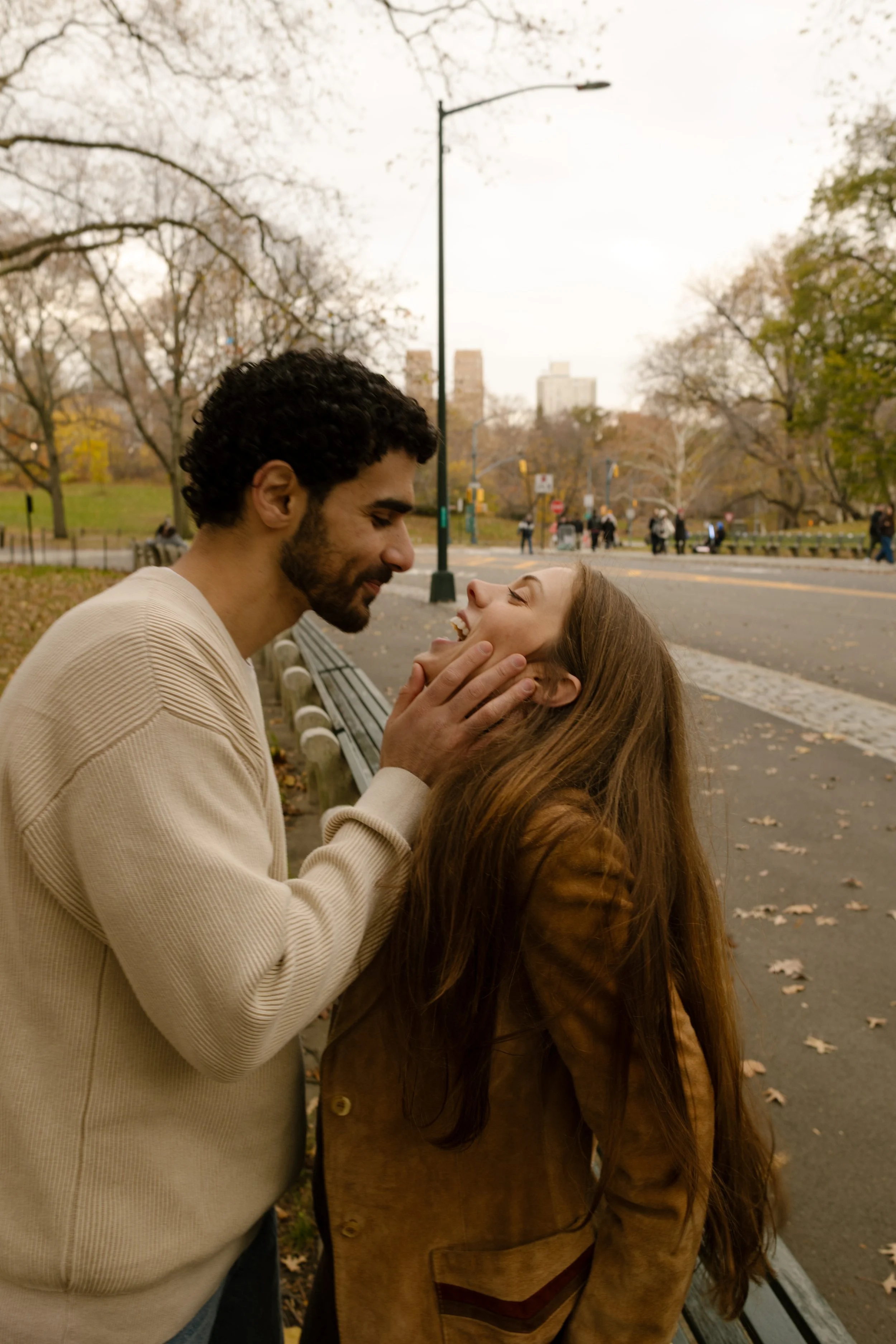 Playful couple sharing a soft moment on a park bench, with New York City in the background.
