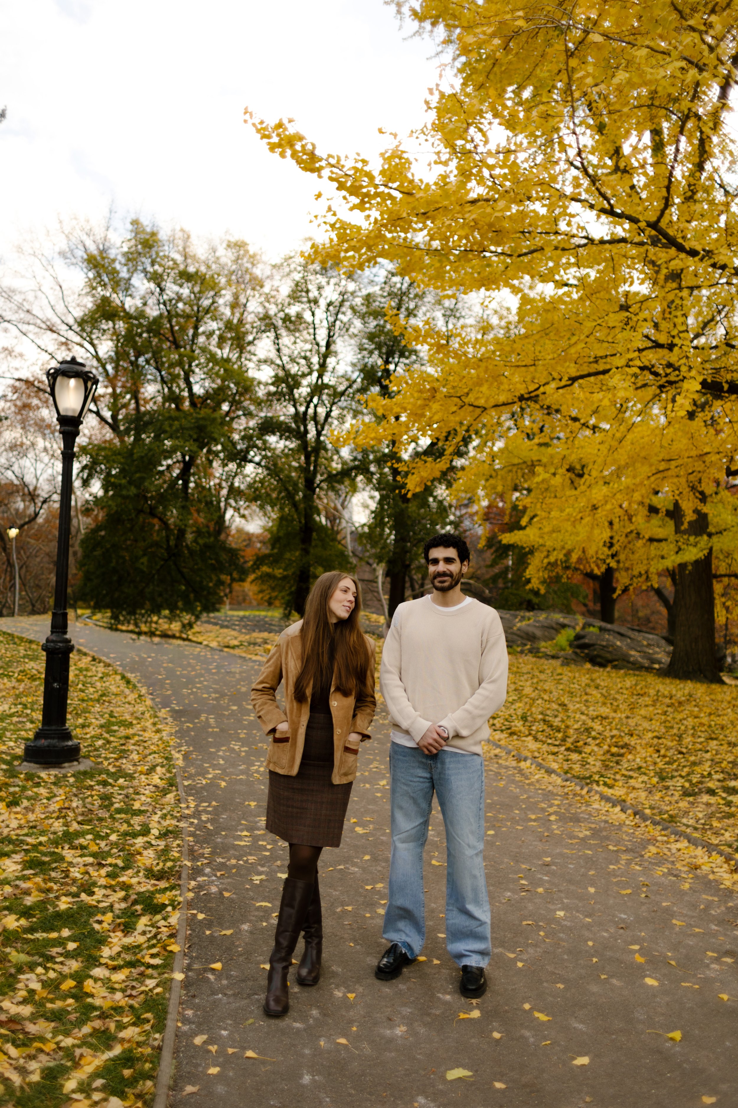 Couple walking a tree-lined path in Central Park surrounded by golden yellow leaves on an overcast fall day.