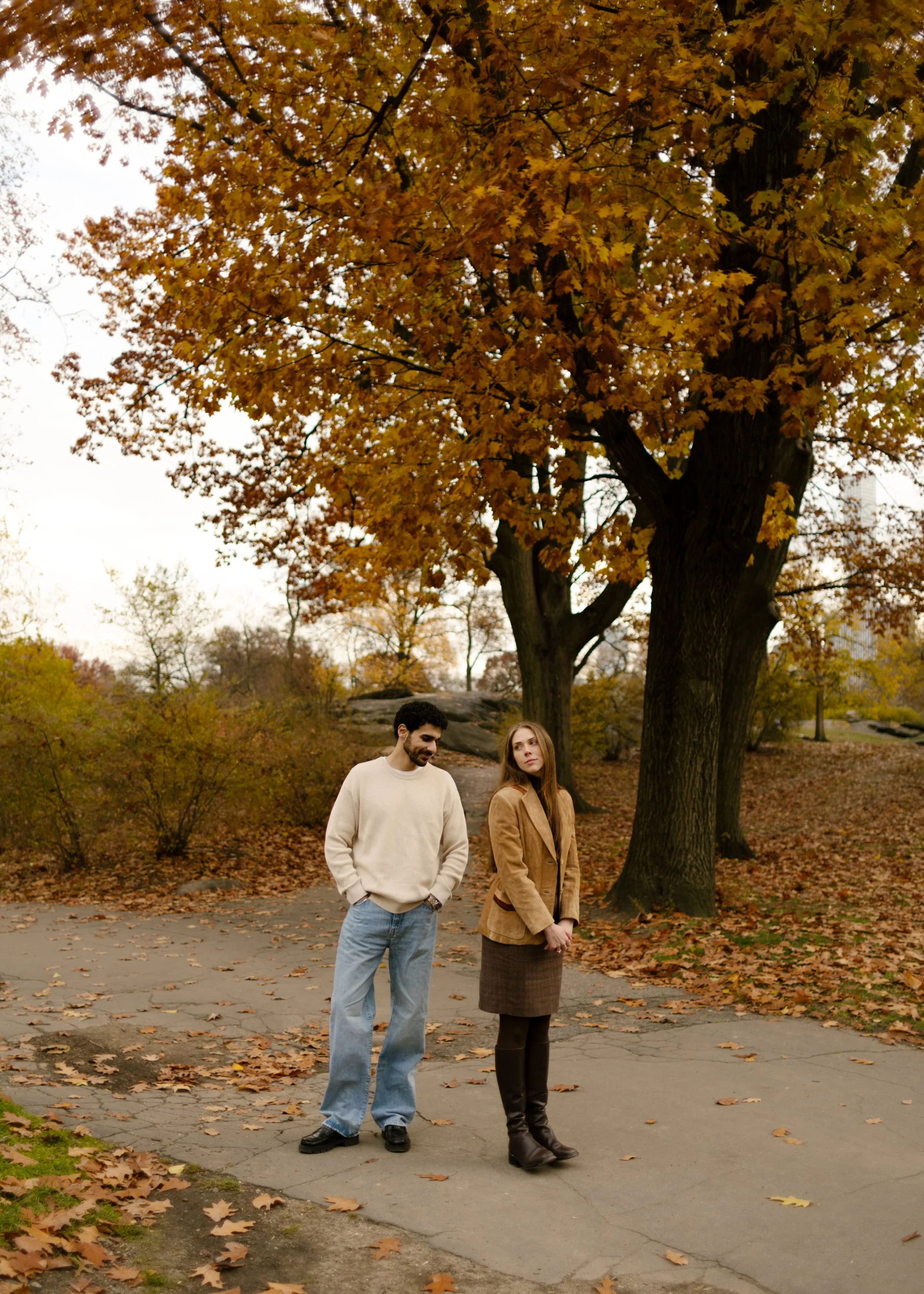 Playful moment between a couple in Central Park, standing apart under a canopy of rust-colored leaves.