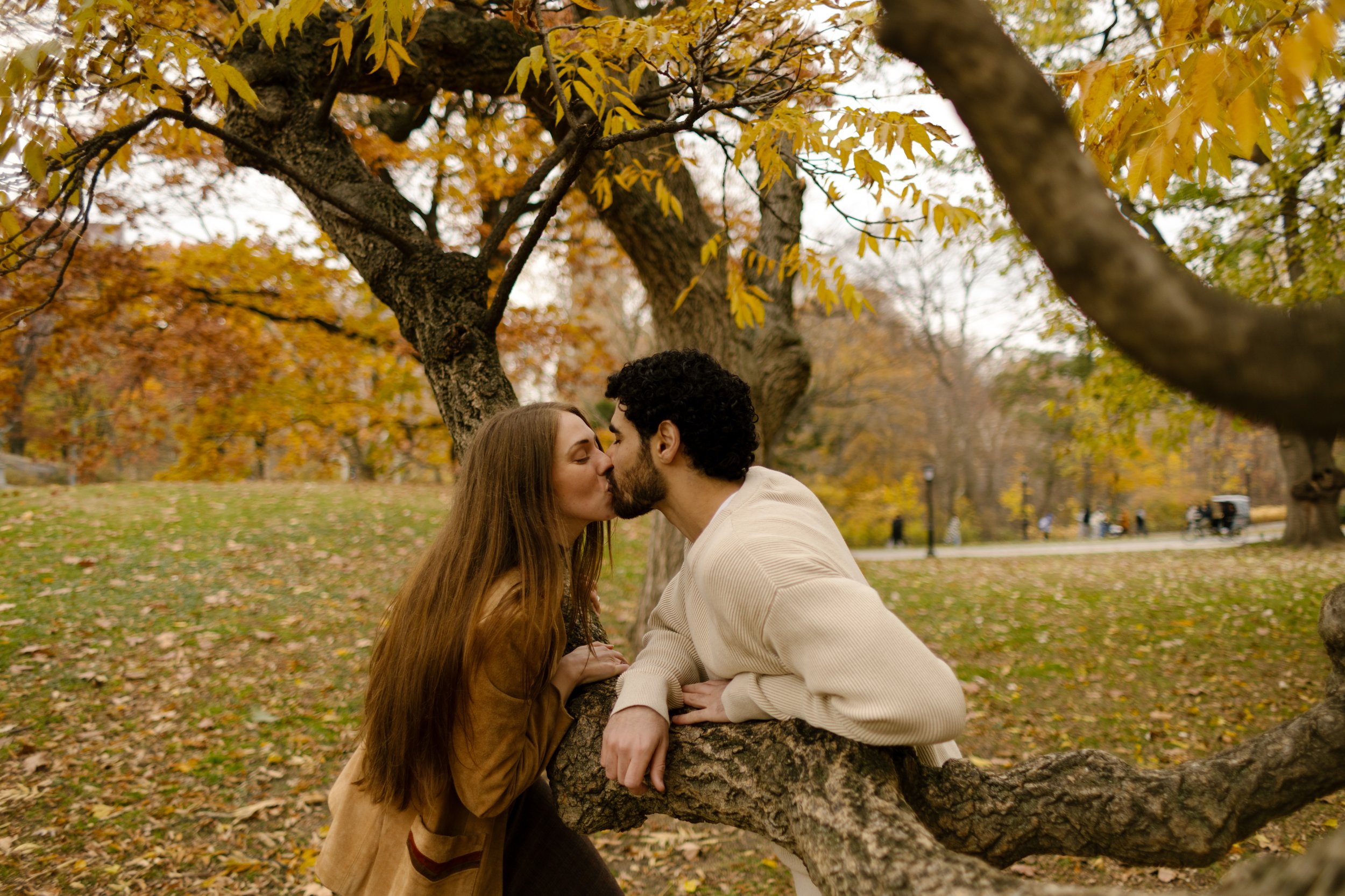 Sweet kiss captured mid-moment between a couple leaning on a twisted tree branch during their Central Park engagement session.