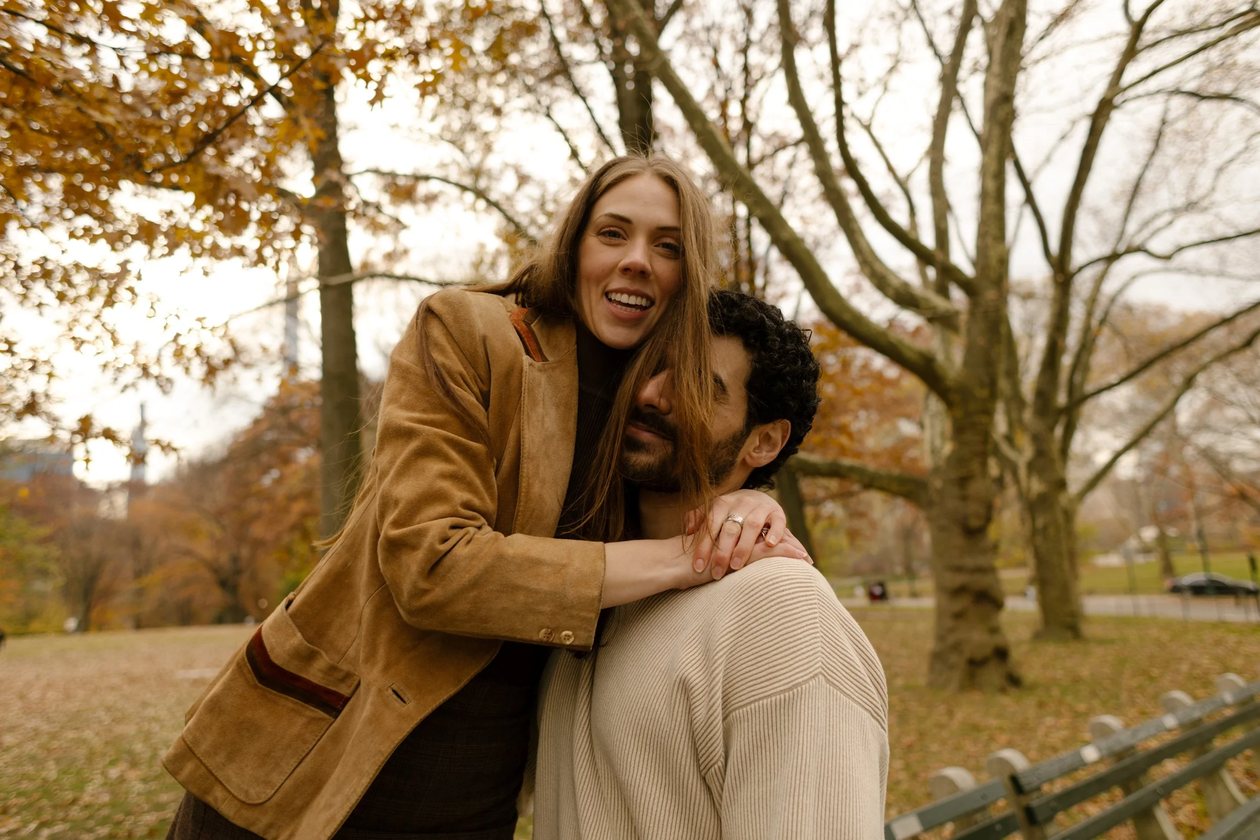 Playful moment as a woman wraps her arms around her partner’s shoulders on a park bench surrounded by fall leaves