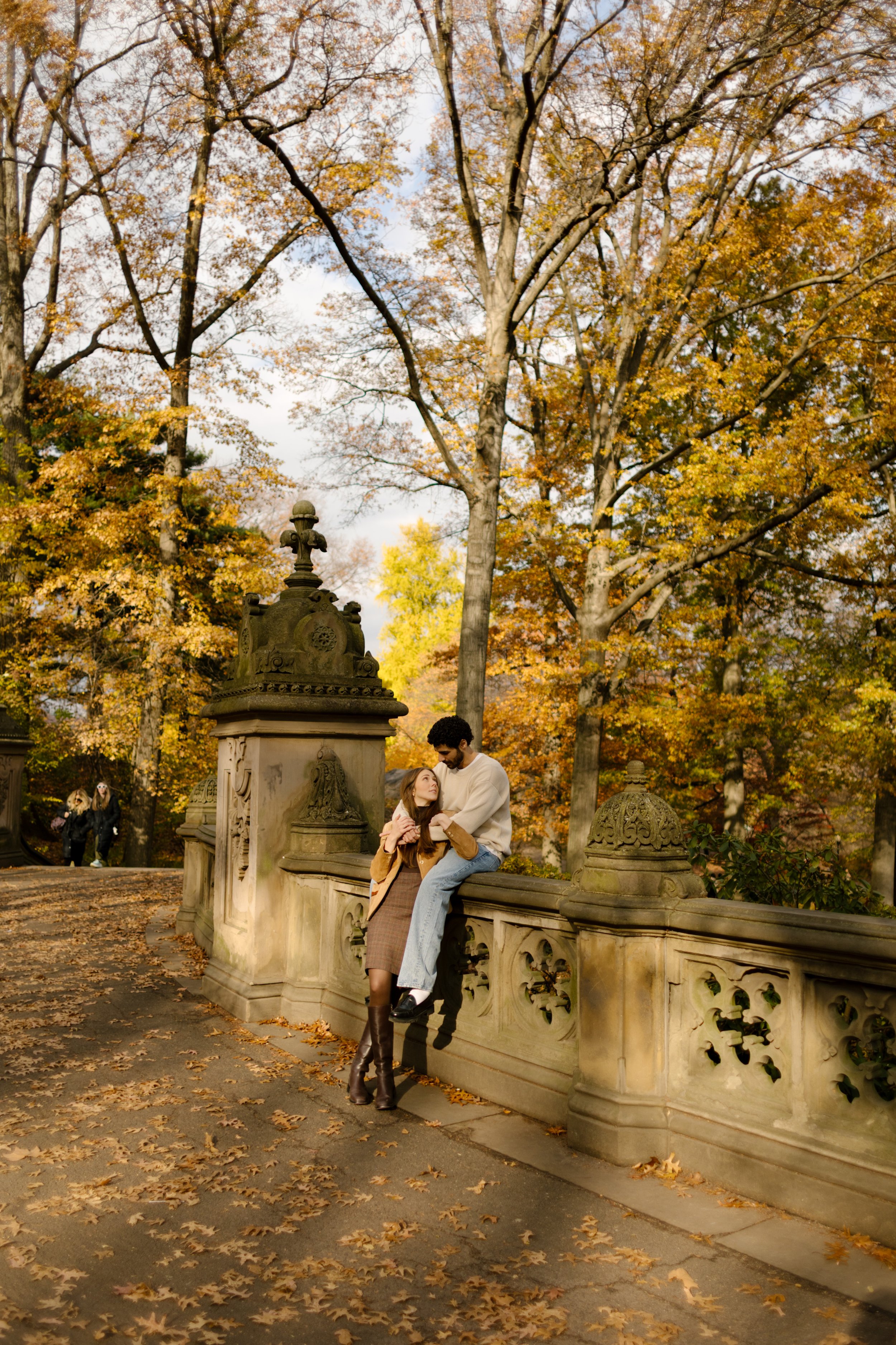 Couple embracing against the Bethesda Terrace railing, surrounded by golden leaves during their Central Park photoshoot.
