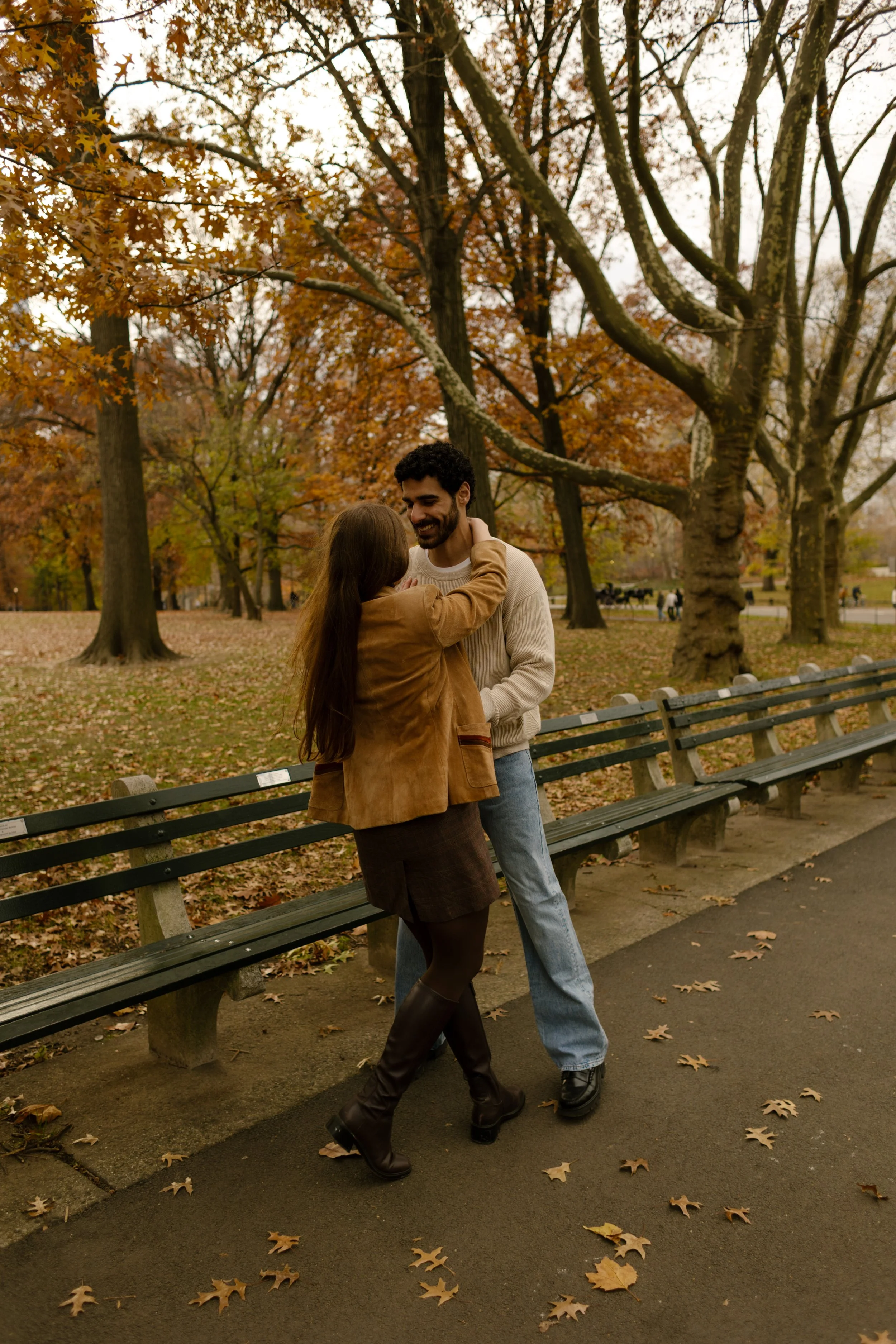 Candid moment of a couple holding each other on the park path with rows of benches and bare fall trees.