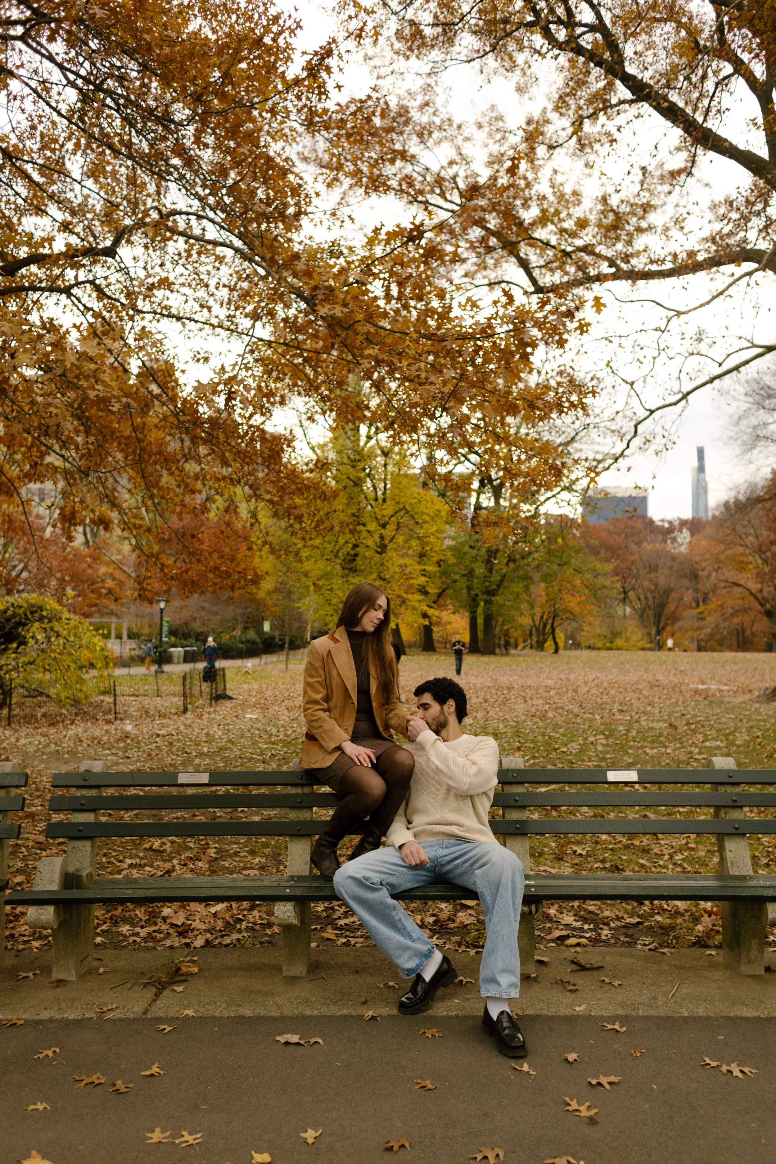 Playful couple on a park bench mid-laugh, with skyscrapers peeking through the golden fall trees of Central Park.