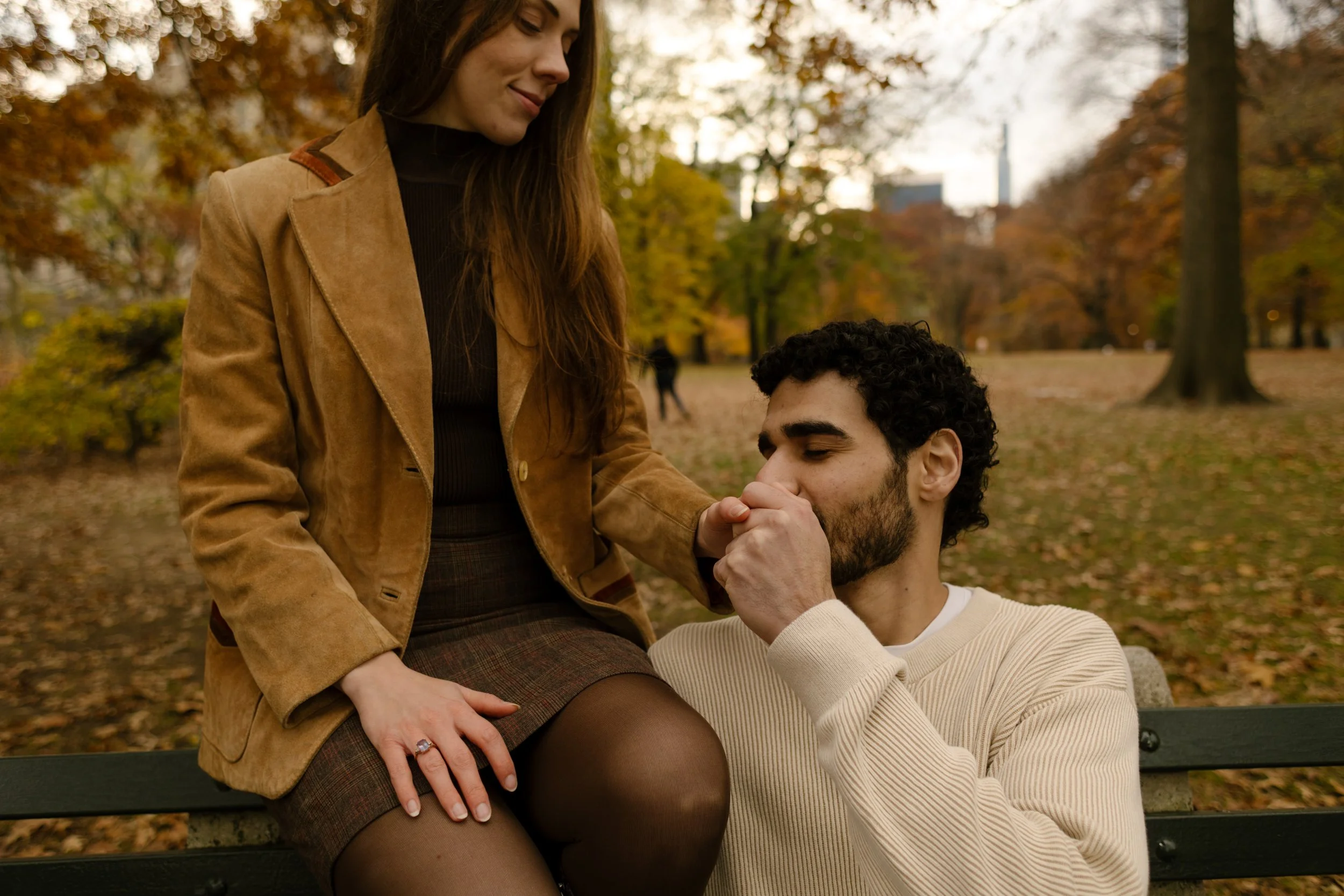 Romantic gesture during a Central Park photoshoot as a man kisses his partner’s hand while she sits on a park bench.
