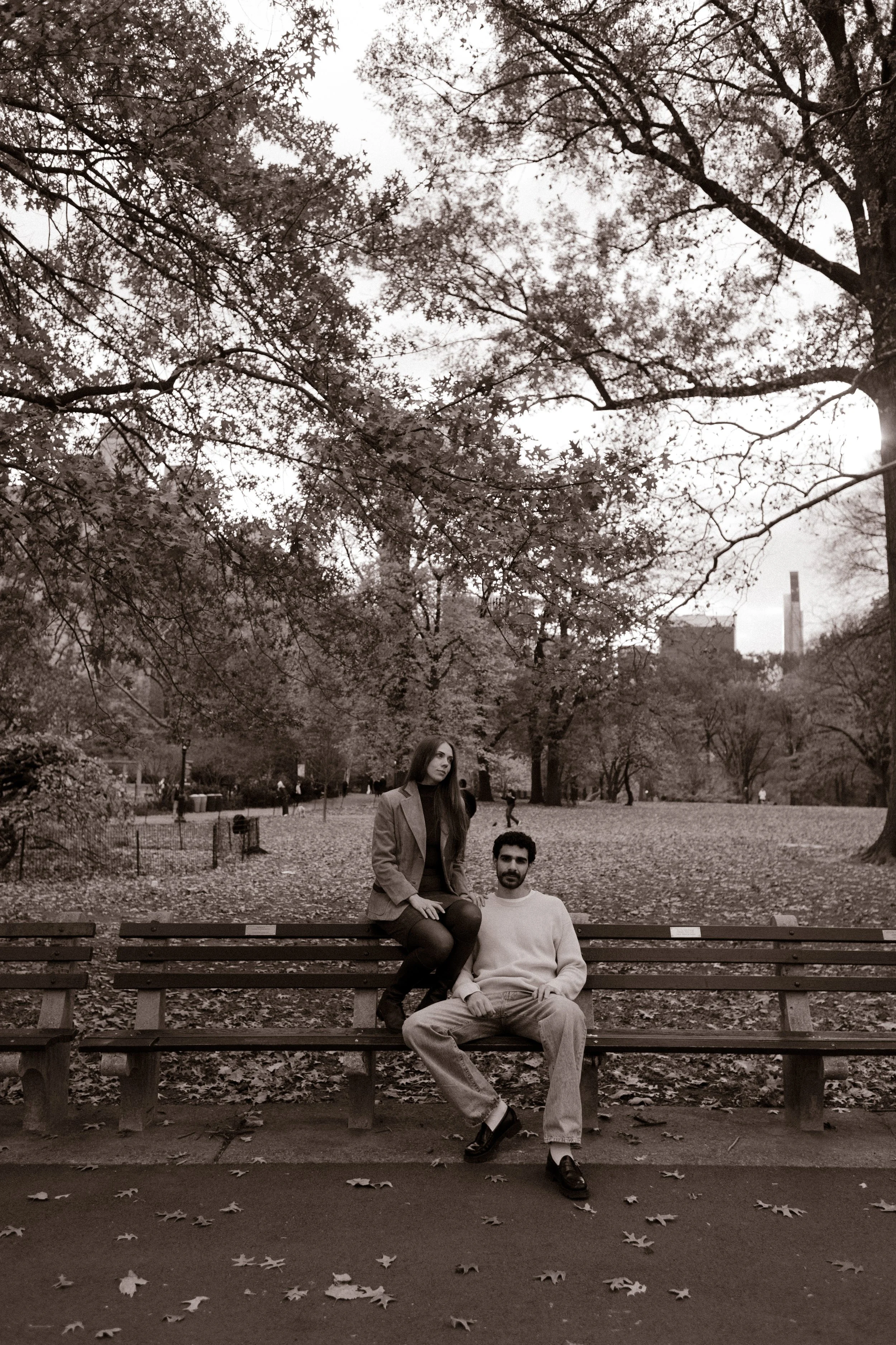 Moody black and white wide shot of a couple sitting on a bench surrounded by tall trees and the city skyline.