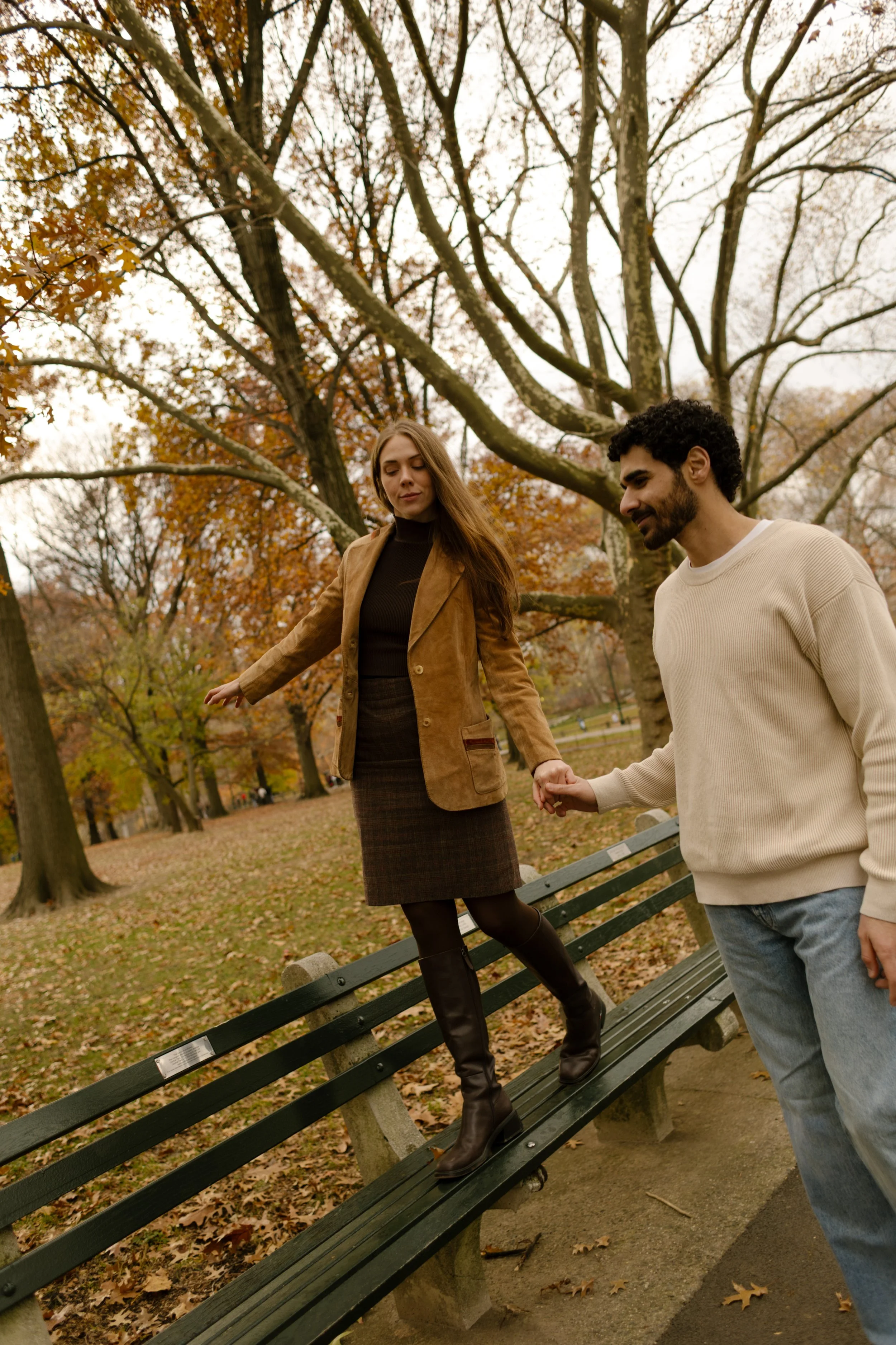 Playful fall moment as a woman walks along a park bench, hand-in-hand with her partner during a Central Park engagement session.
