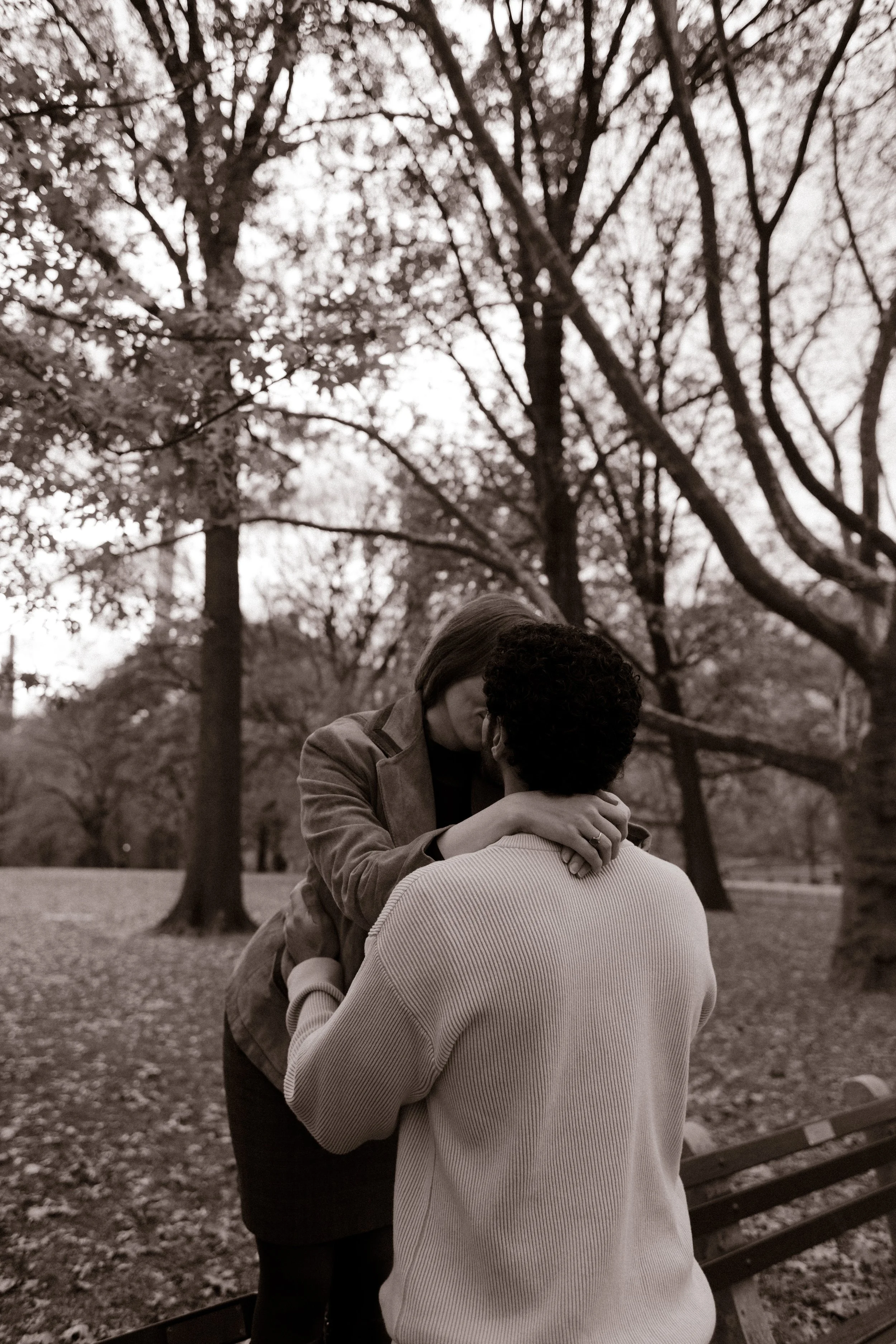 Black and white photo of a couple kissing on a park bench during their intimate Central Park photoshoot, surrounded by bare winter trees.