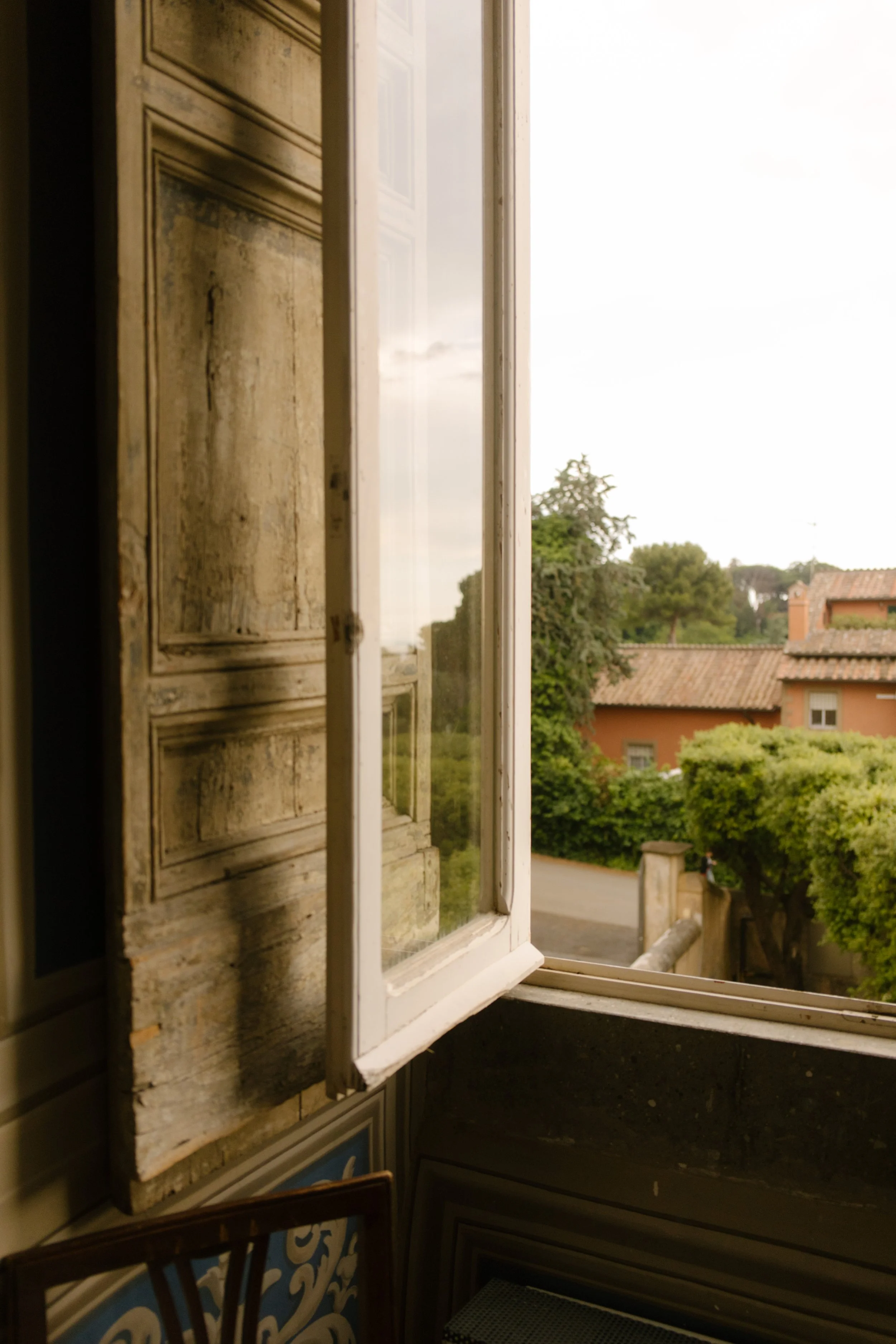 Window is open overlooking Italian vineyard with tall trees and historic buildings.