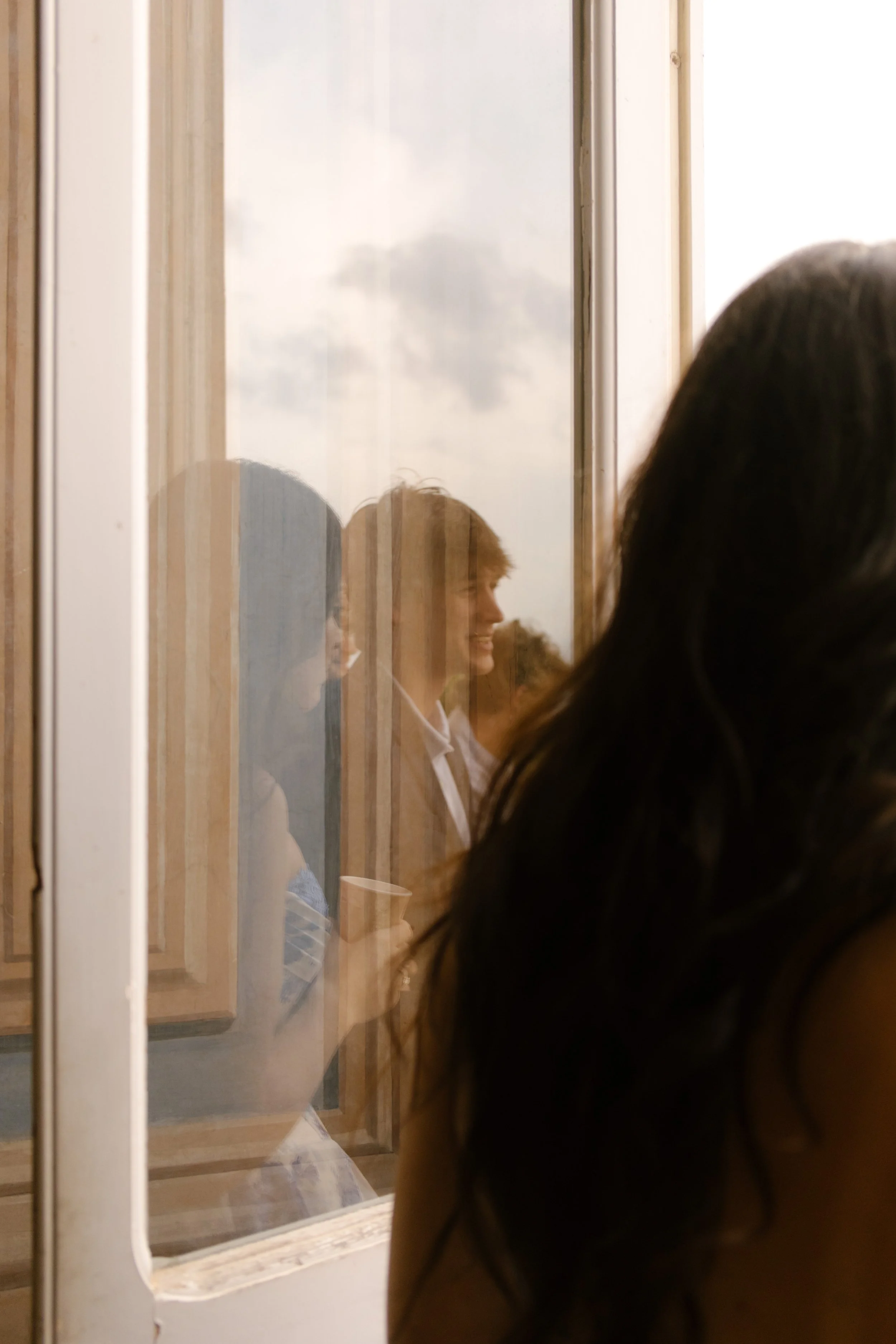 A soft reflection of the couple captured through a glass door, the bride’s silhouette in the foreground as the groom smiles faintly. An intimate, almost-secret moment during a wedding in Italy.