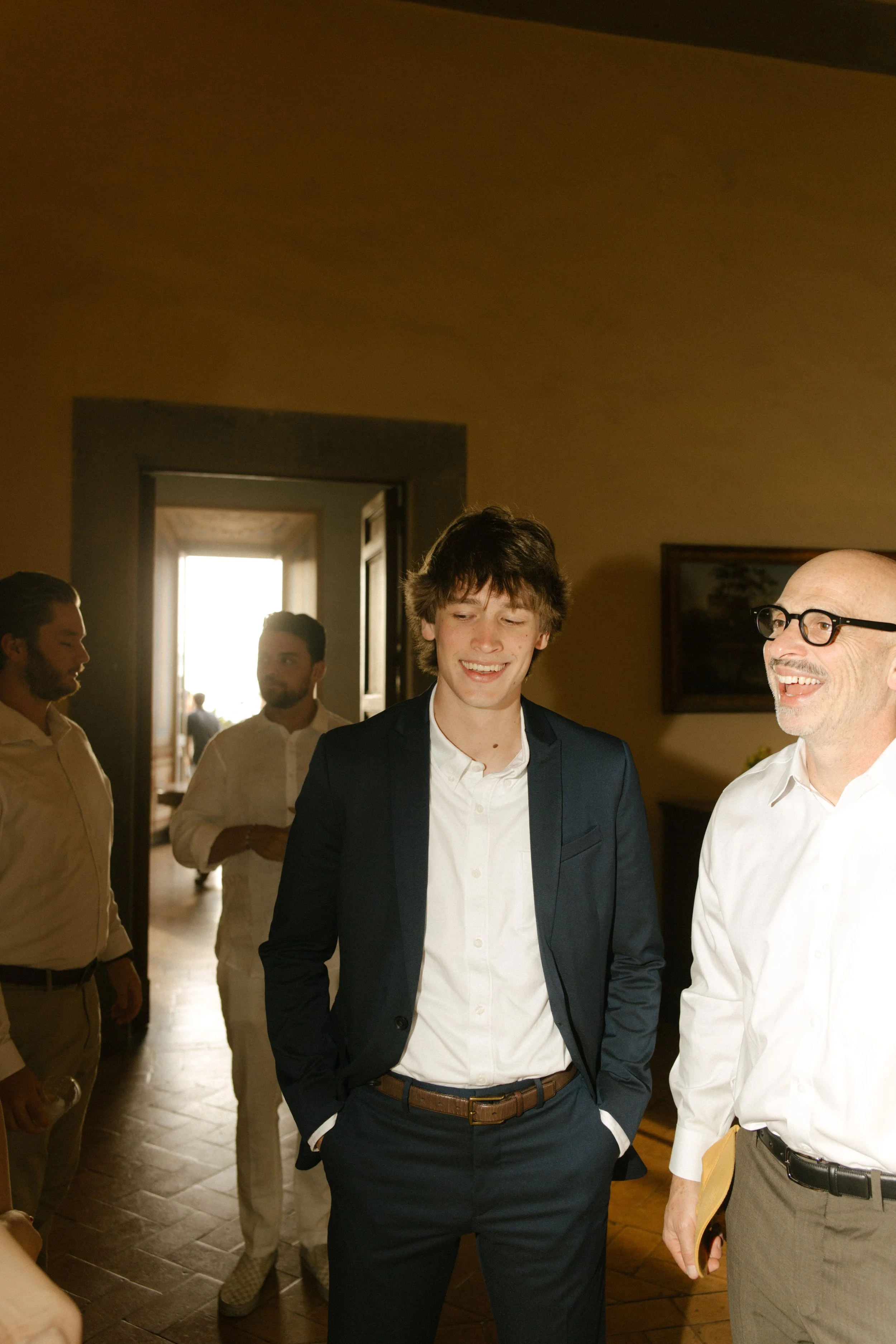 The groom laughs with guests in a sunlit villa hallway, hands in pockets and joy written all over his face. Golden hour banter and pre-ceremony buzz.