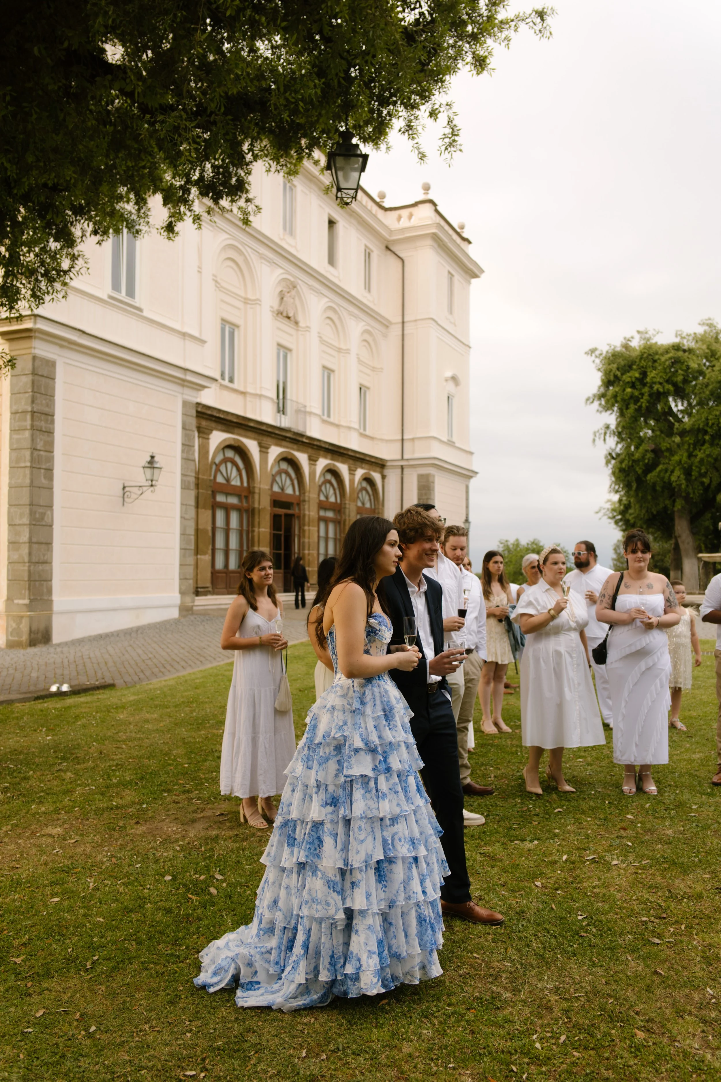 The bride and groom stand together as guests gather nearby on the villa lawn, champagne flutes raised and dresses flowing. Surrounded by love at a wedding in Italy.