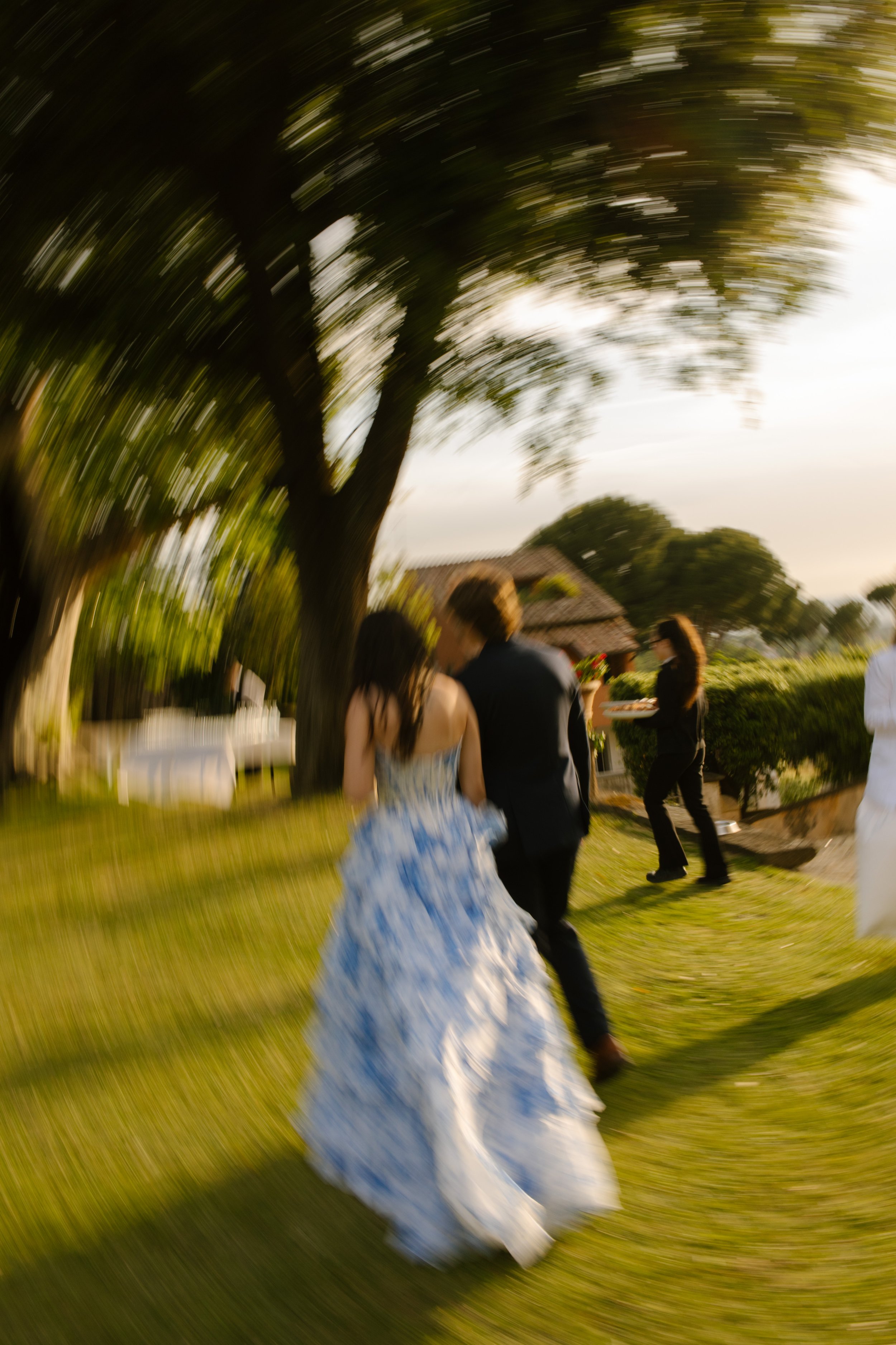 A dreamy motion blur of the couple walking hand-in-hand across the lawn, sunlight streaking through the trees. Running headfirst into forever, slightly out of focus.