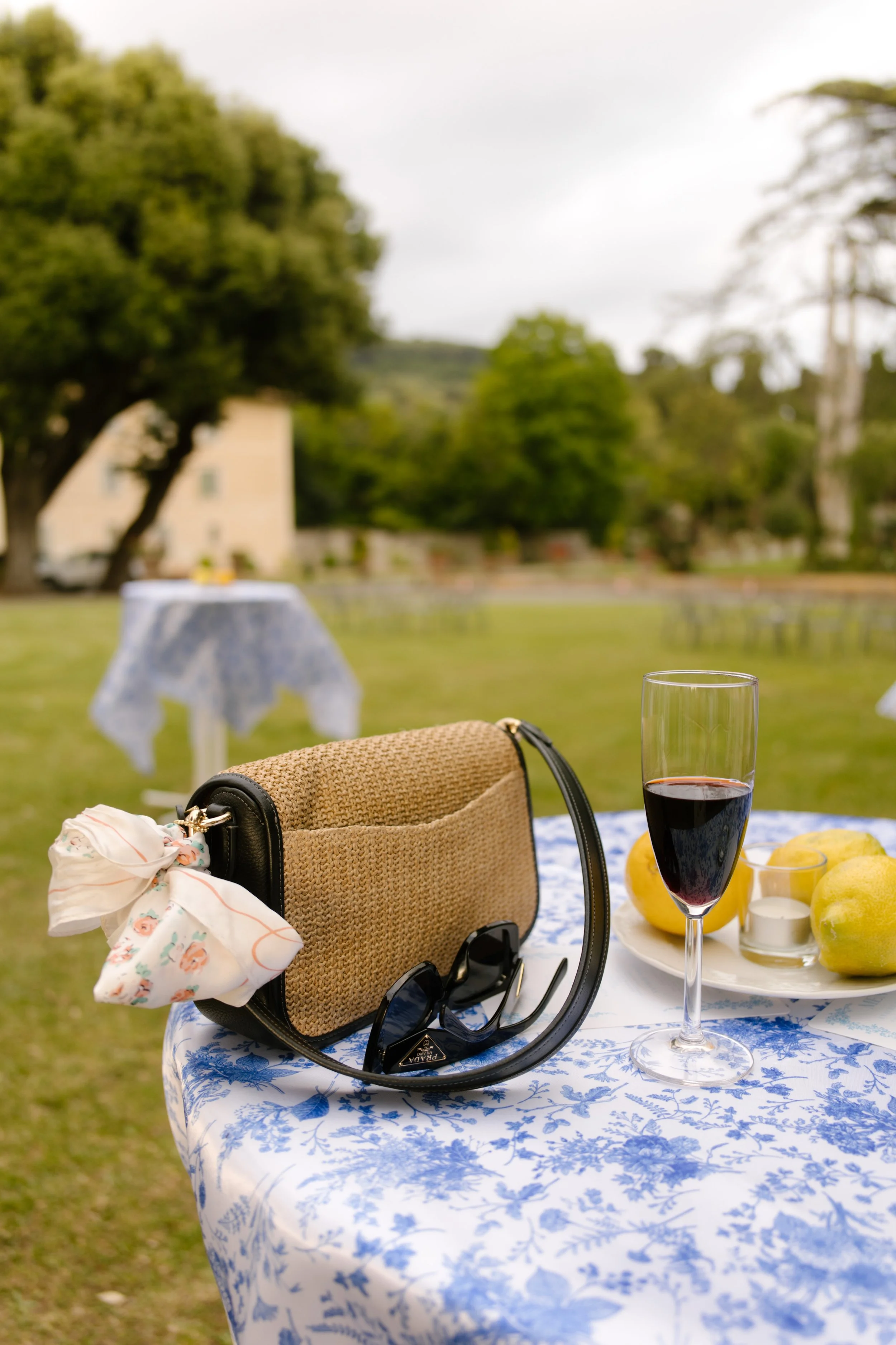 A straw bag, sunglasses, and glass of wine rest on a blue toile tablecloth next to a bundle of lemons. Still life meets storybook at a wedding in Italy.