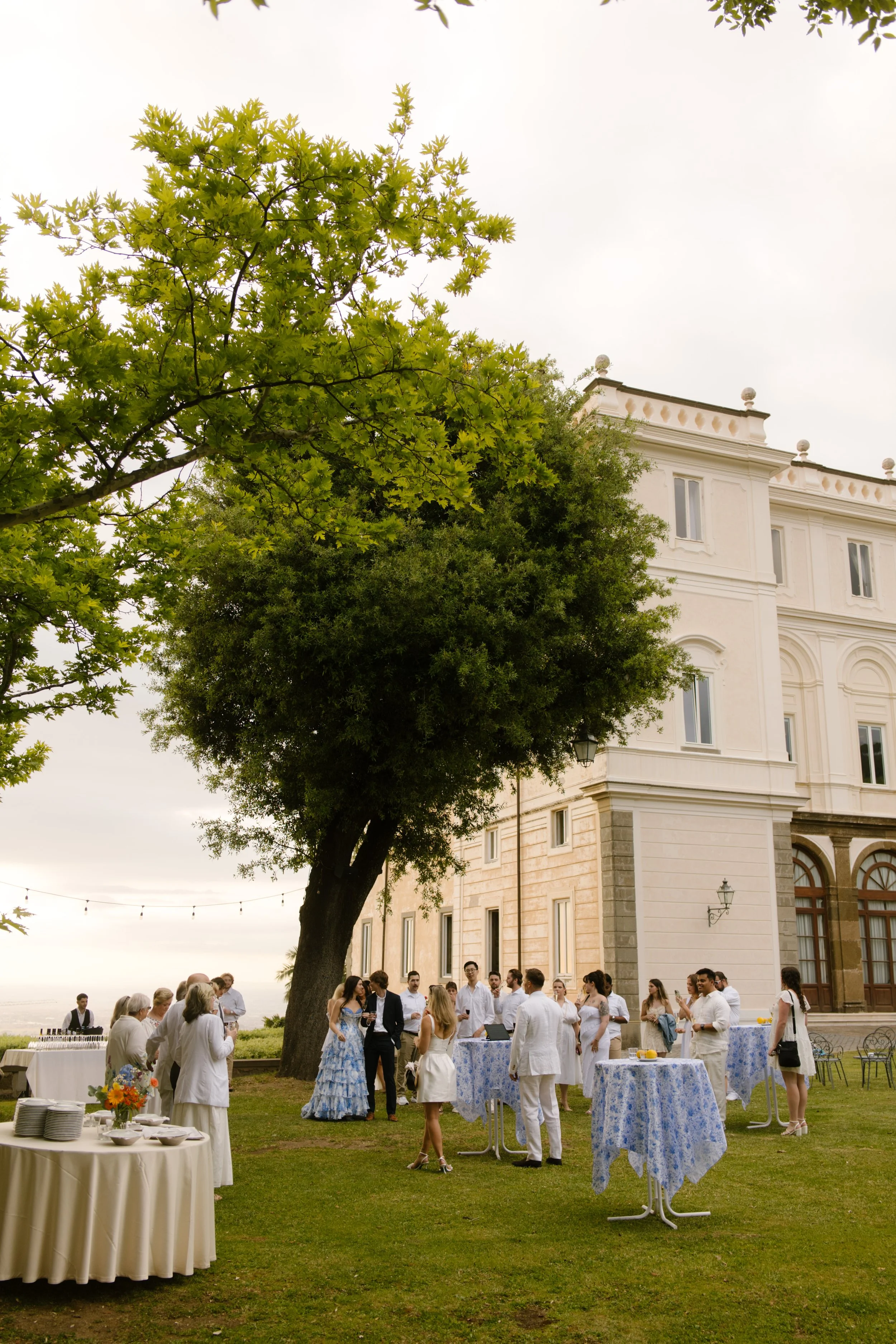 Guests mingle under a sprawling tree on the lawn of a grand Italian villa, blue and white linens fluttering in the breeze. Lawnside mingling at a wedding in Italy.