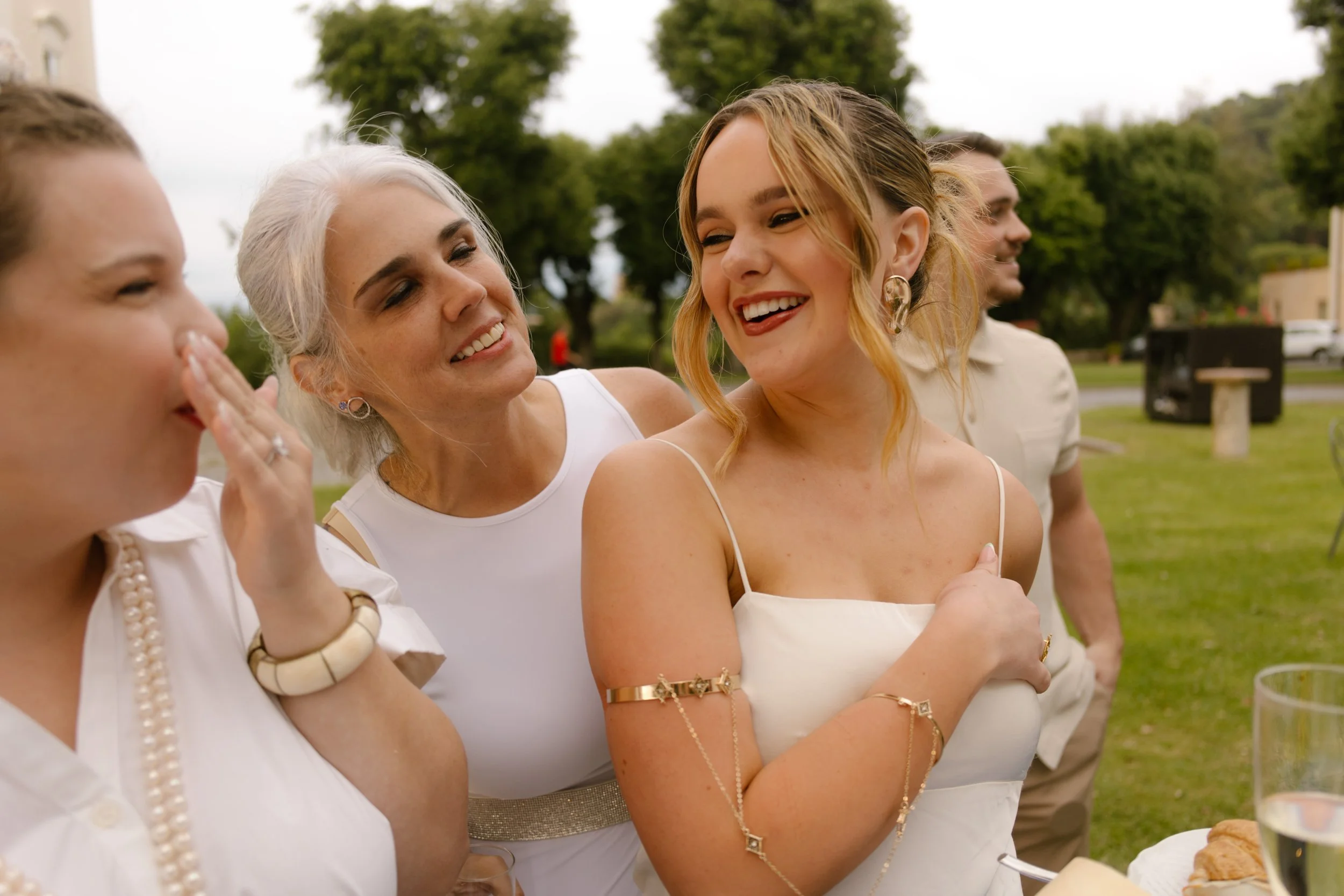 A woman in a white slip dress beams as she chats with loved ones, laughter spilling from the group. Candid connection during a wedding in Italy.