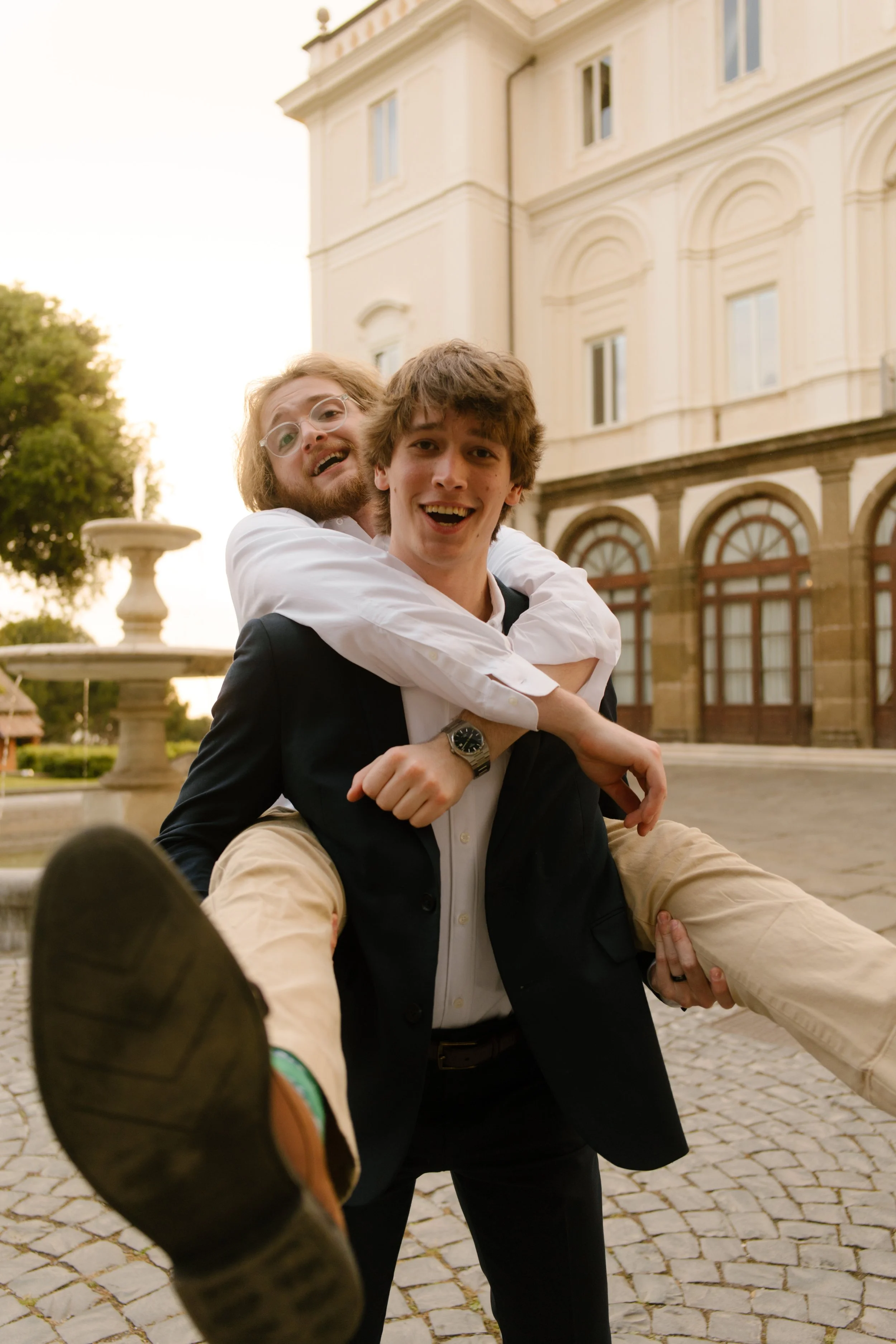 Two groomsmen playfully goof off in front of the villa, one giving the other a piggyback ride with joyful chaos in the air. Pre-ceremony antics full of energy and friendship.
