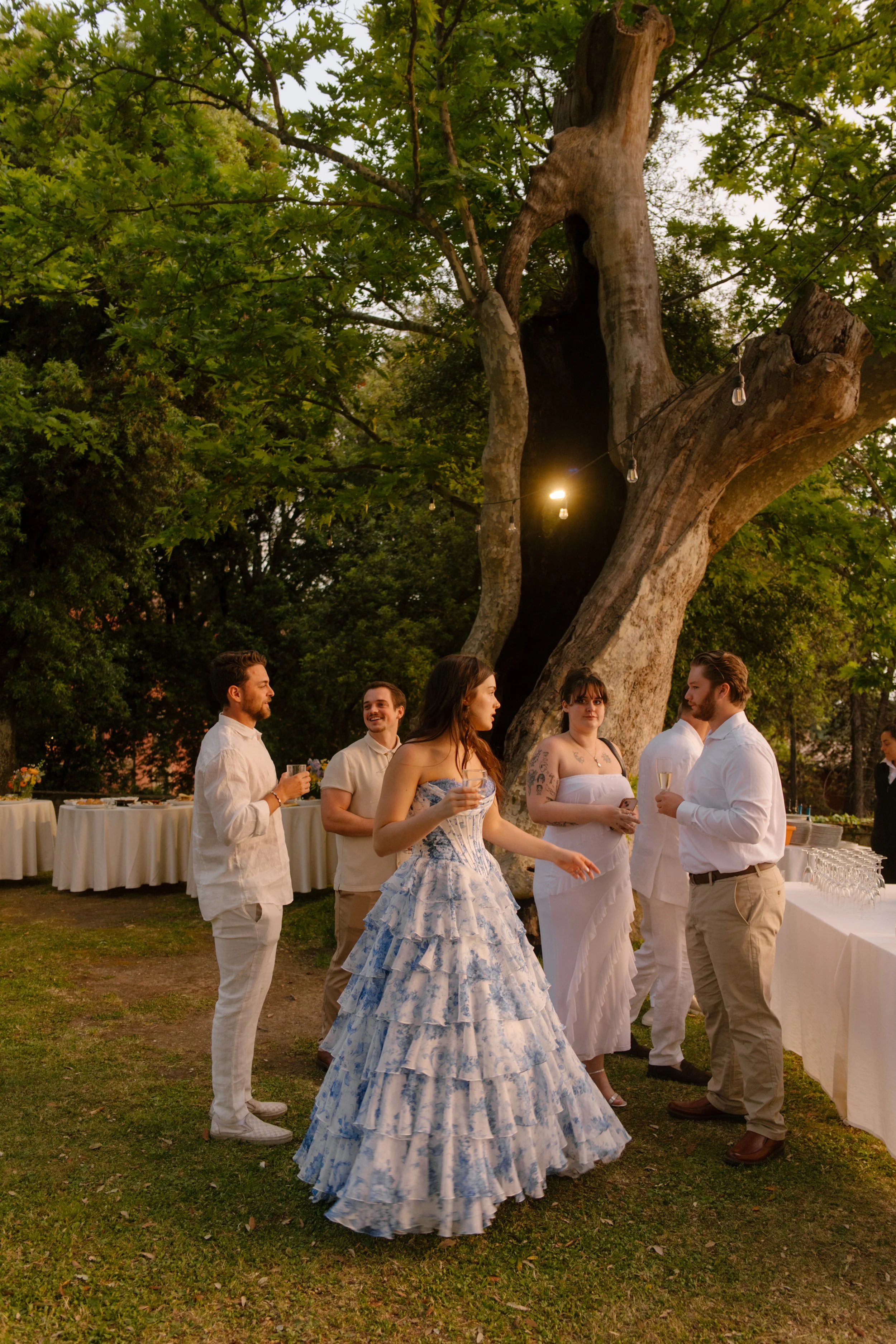 The bride in her blue ruffled gown gestures animatedly as she chats with guests under a towering tree wrapped in string lights. Golden hour conversations that feel like home.