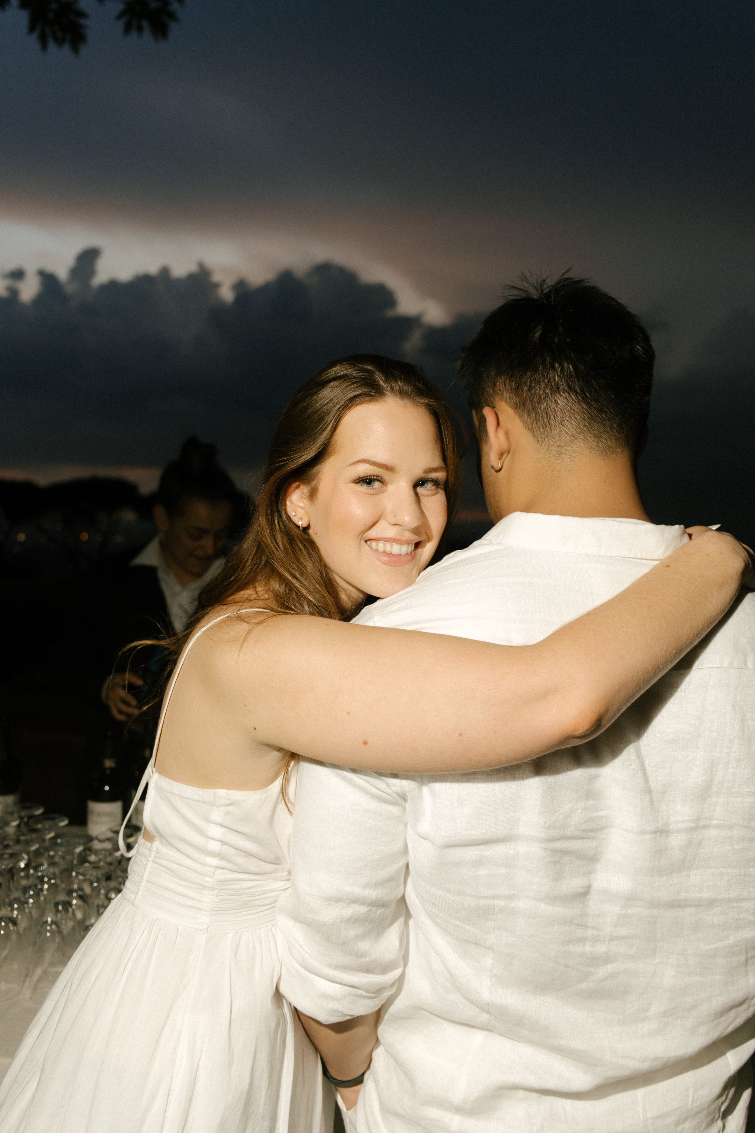A woman in white smiles over her shoulder with her arm wrapped around her partner, champagne flutes and stormy skies in the background. An intimate reception moment full of warmth and weather.