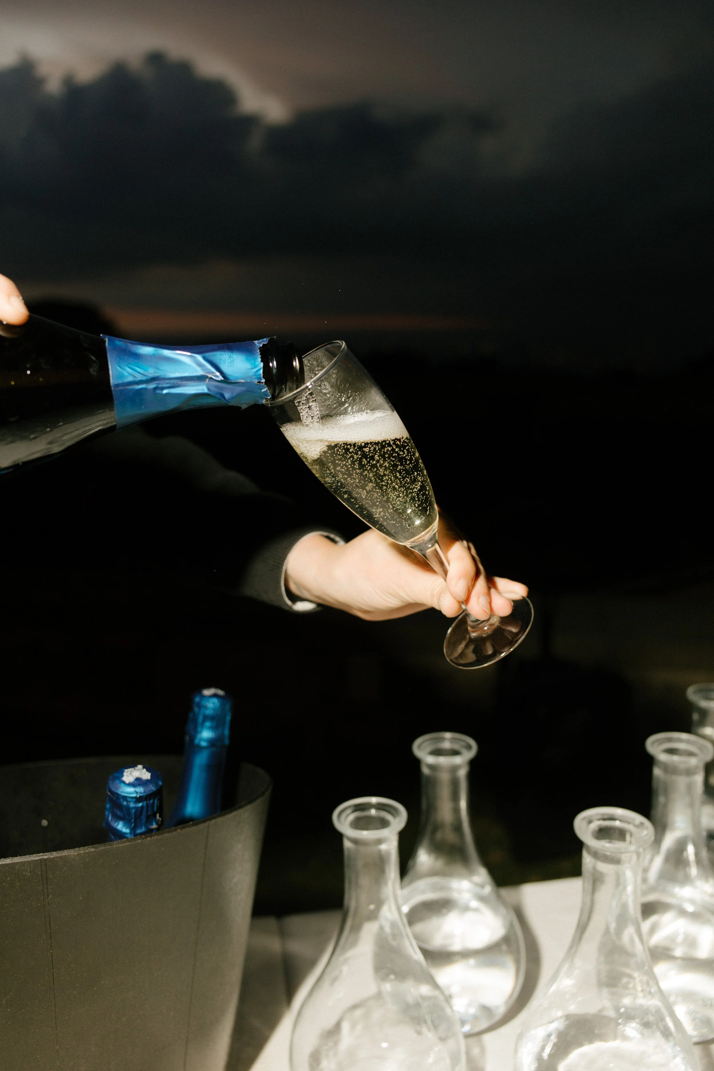 Champagne is poured into a glass as night falls, surrounded by empty carafes and anticipation. Celebration in full swing at a wedding in Italy.