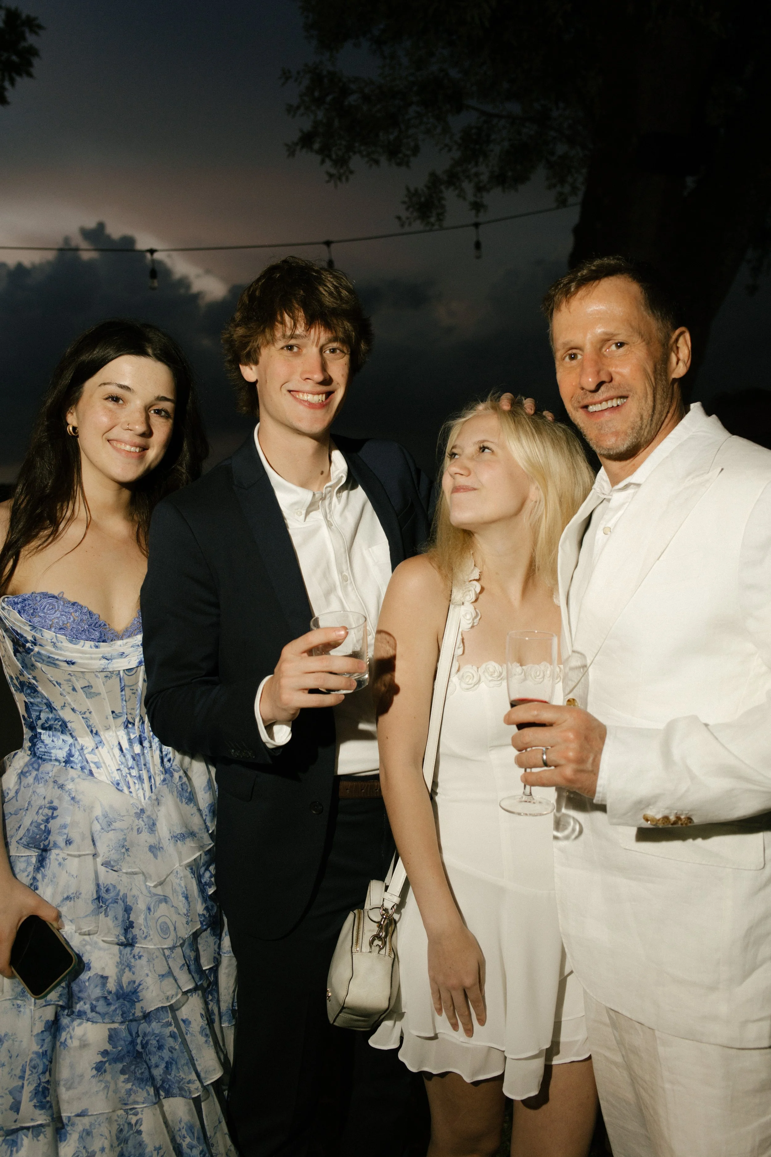The bride and groom pose with two guests dressed in white, everyone glowing under string lights and evening sky. Family and friends gathered for a wedding in Italy.