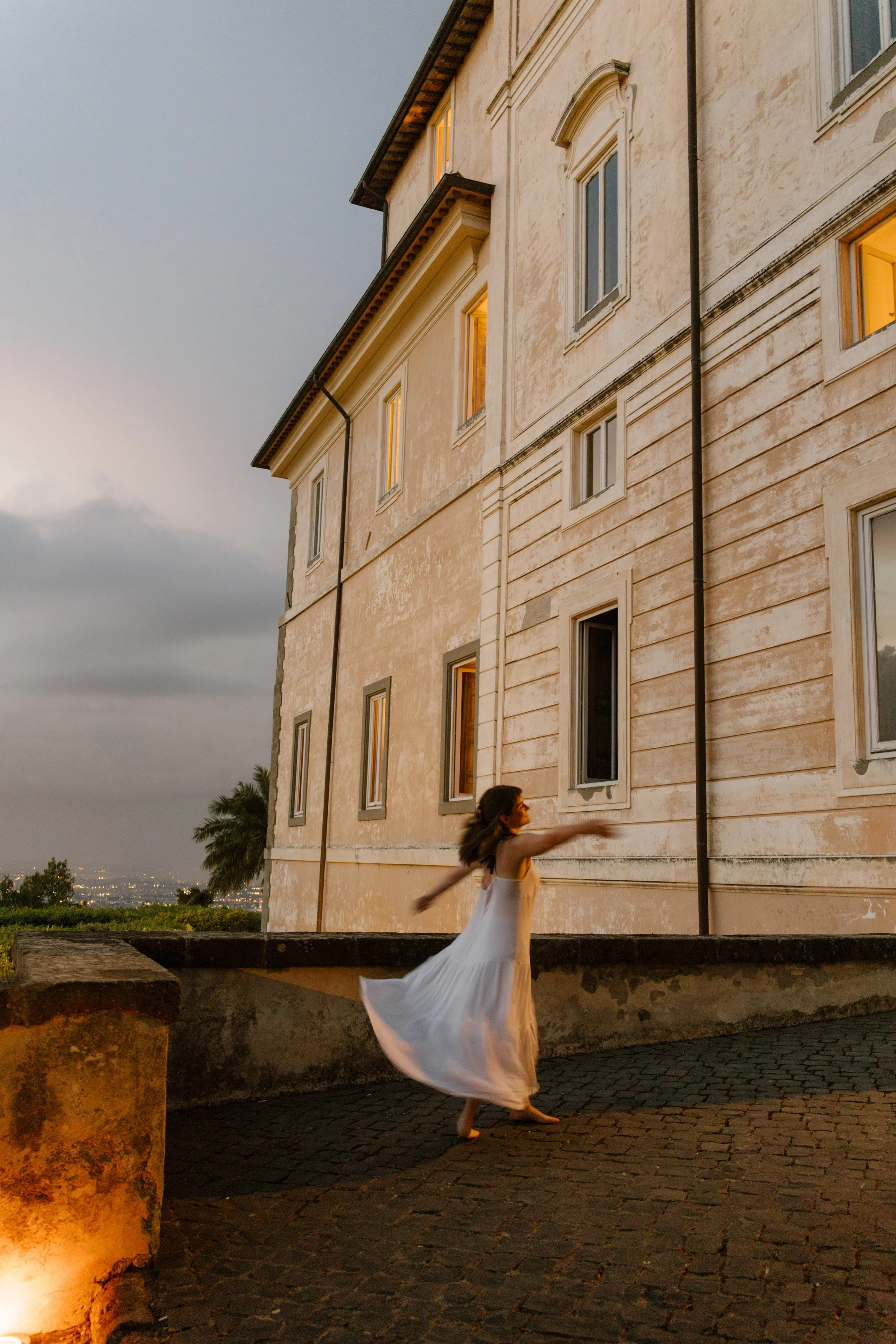 A guest in a flowing white dress twirls beneath moody skies beside a historic villa. Playful joy and soft movement captured before the rain.