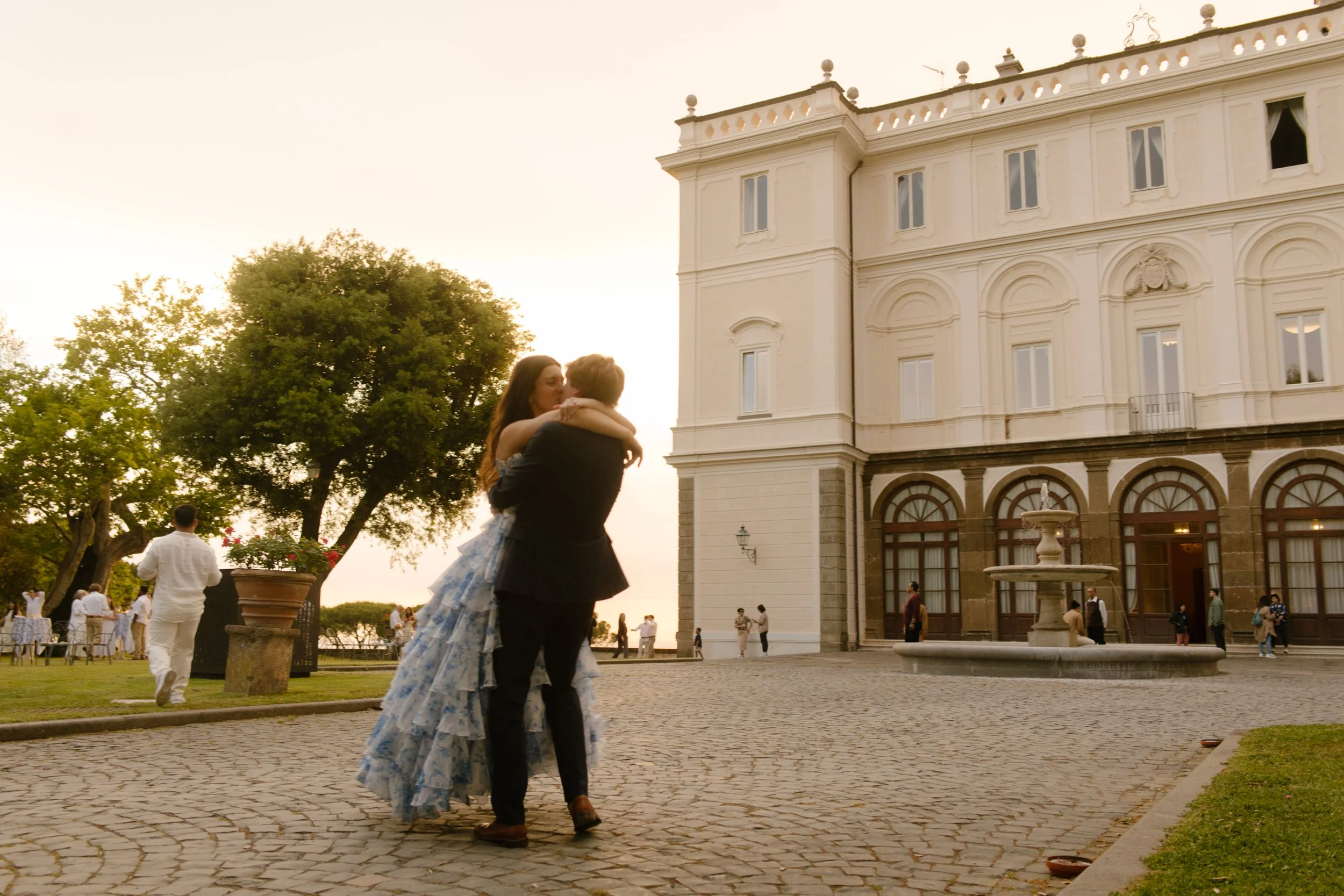 The groom lifts the bride in his arms for a quiet embrace on the cobblestone drive, golden light pouring over the villa behind them. Dreamy golden hour moment during a wedding in Italy.