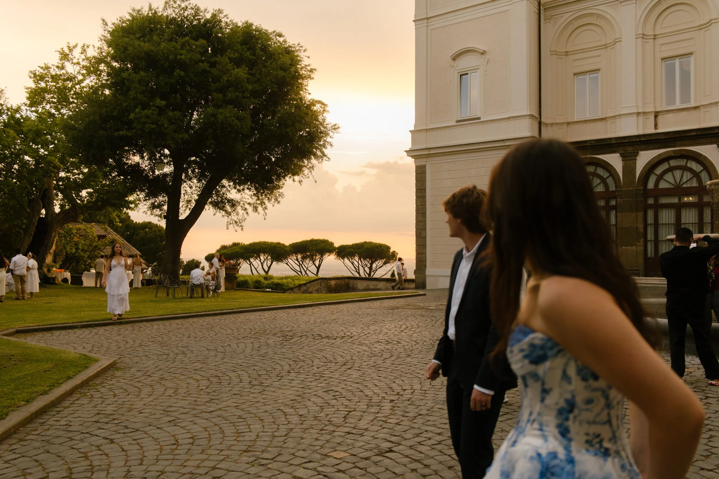 The bride and groom glance toward the lawn as golden hour stretches over the sea, guests scattered beneath trees. Looking out at love unfolding during a wedding in Italy.