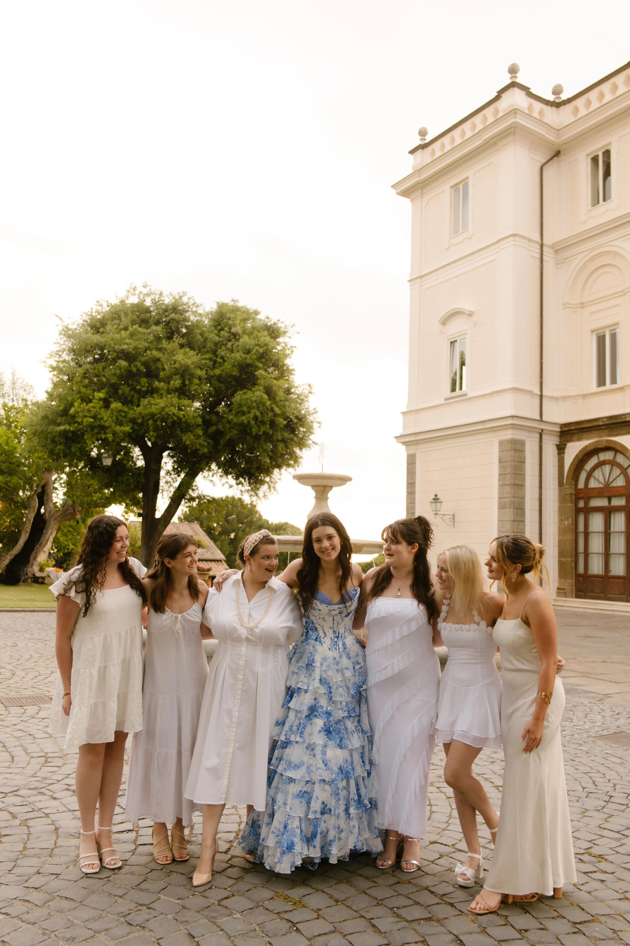 A bride in a blue and white gown stands surrounded by her bridesmaids in soft neutrals, laughing together in front of a historic Italian villa. Wedding in Italy vibes with major main character energy.