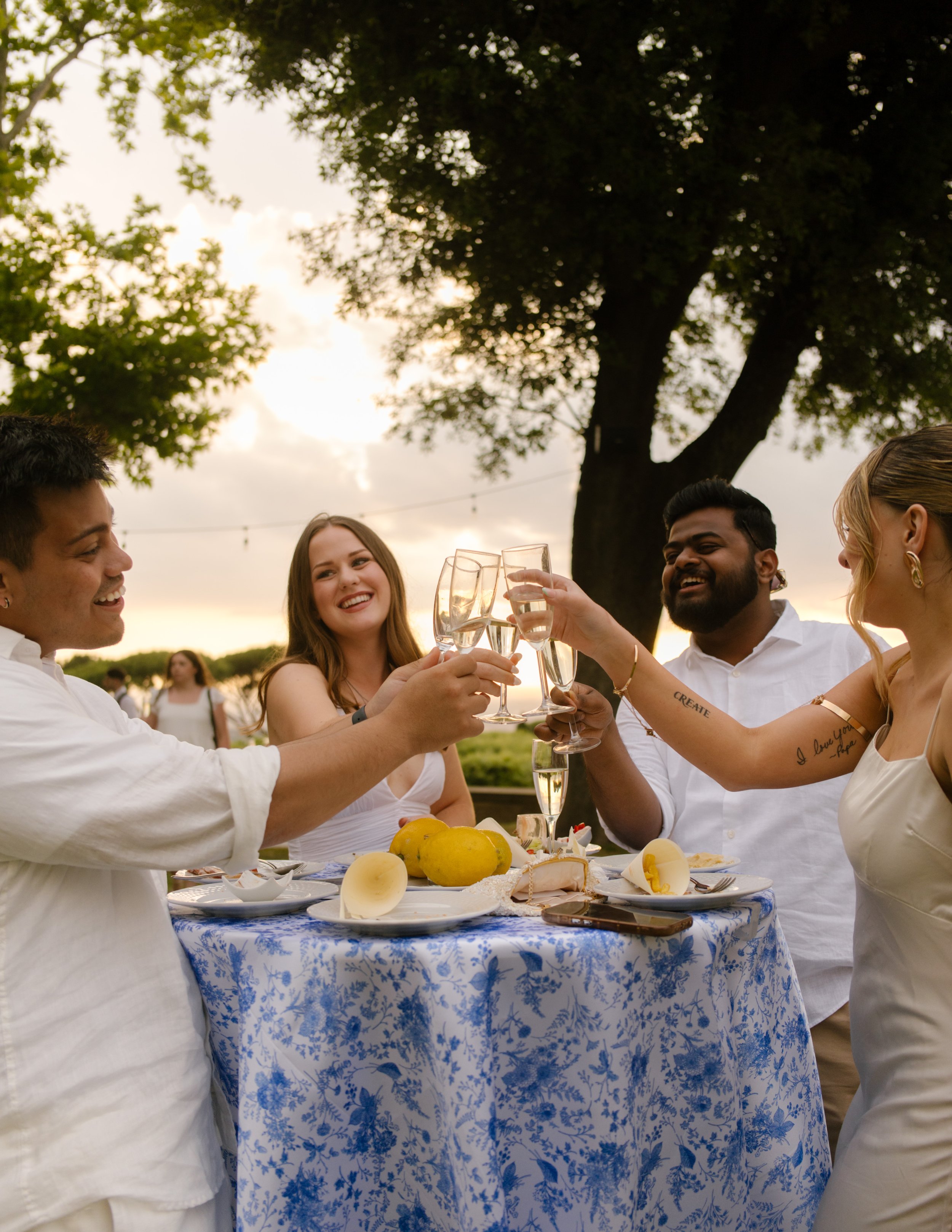 Four friends clink glasses at a blue and white table, mid-toast during golden hour, surrounded by lemon slices and joy. A candid, sun-soaked dinner moment under the Italian sky.