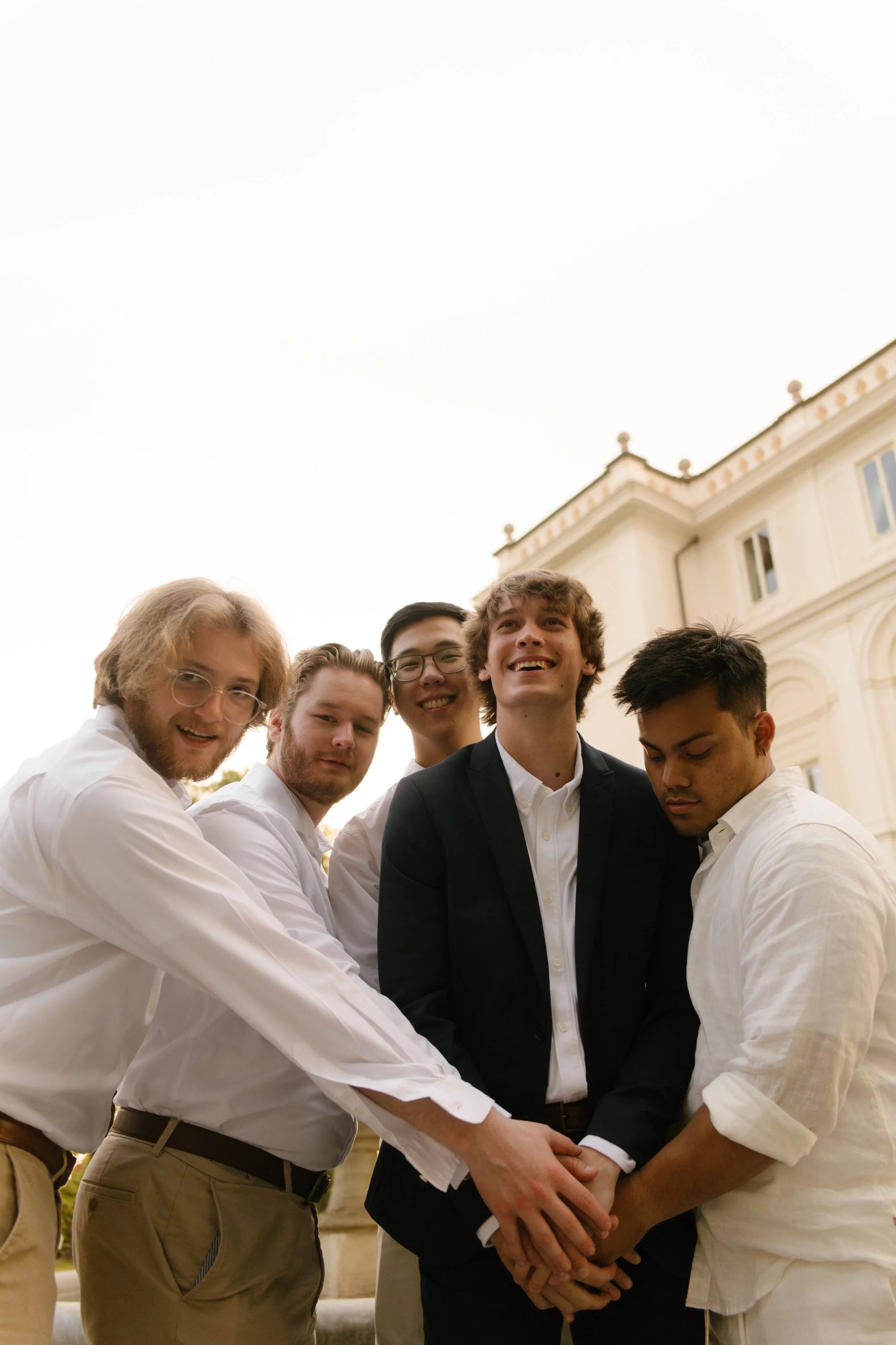 A groom and his groomsmen stack their hands together, beaming in front of an elegant villa. Playful pre-wedding energy captured before the ceremony in Italy.