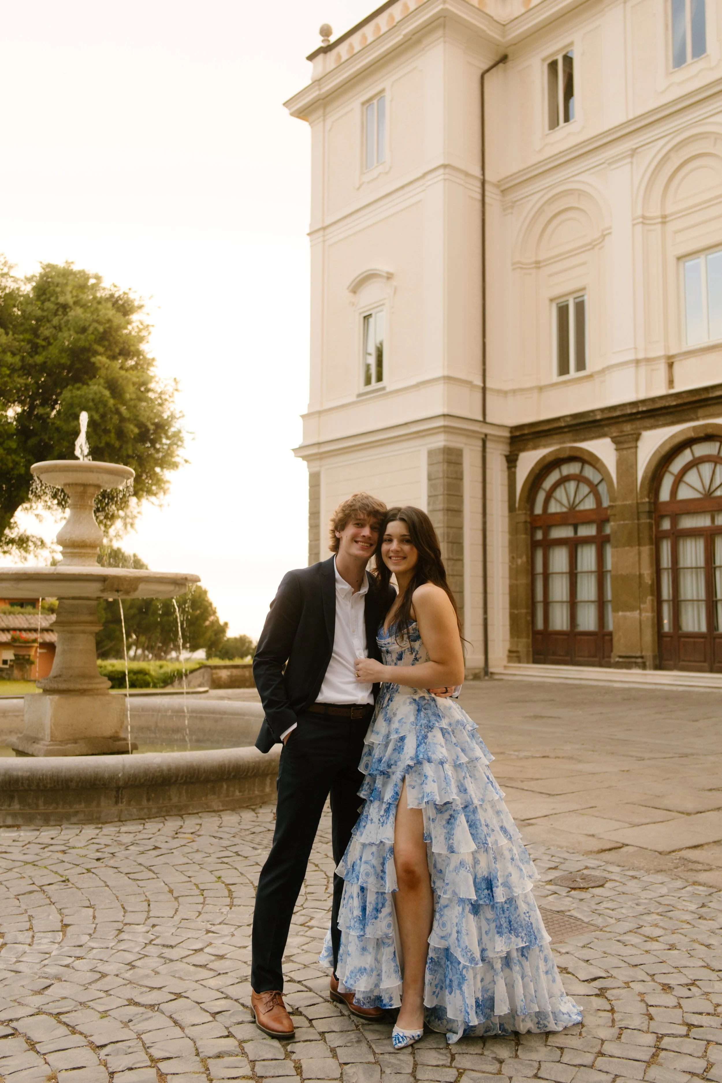 The couple shares a quiet moment in front of the villa fountain, dressed in a sleek suit and flowing floral gown. Romantic sunset portraits during a wedding in Italy.