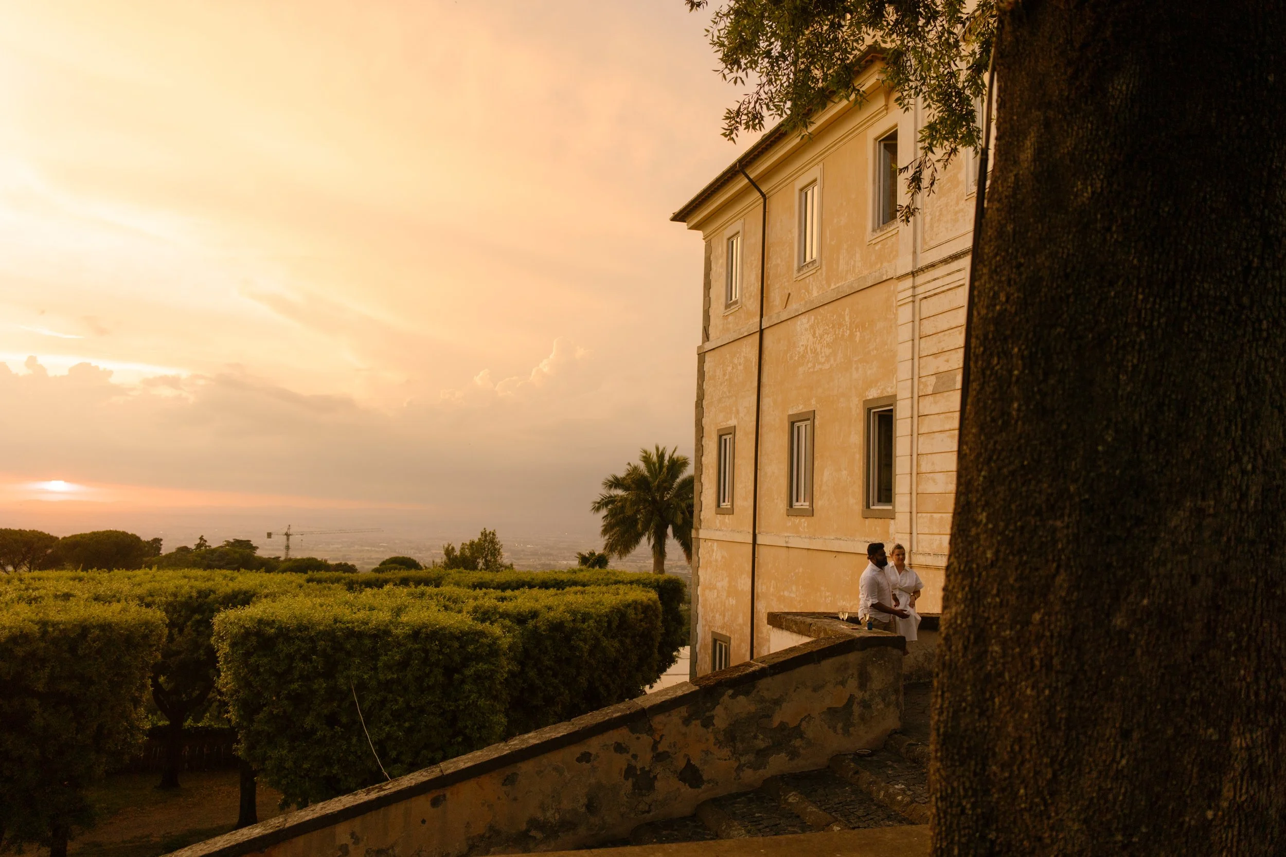 A couple sits tucked into a stone ledge outside the villa as the sky glows in soft amber tones. Golden hour stillness and stolen kisses after the chaos.