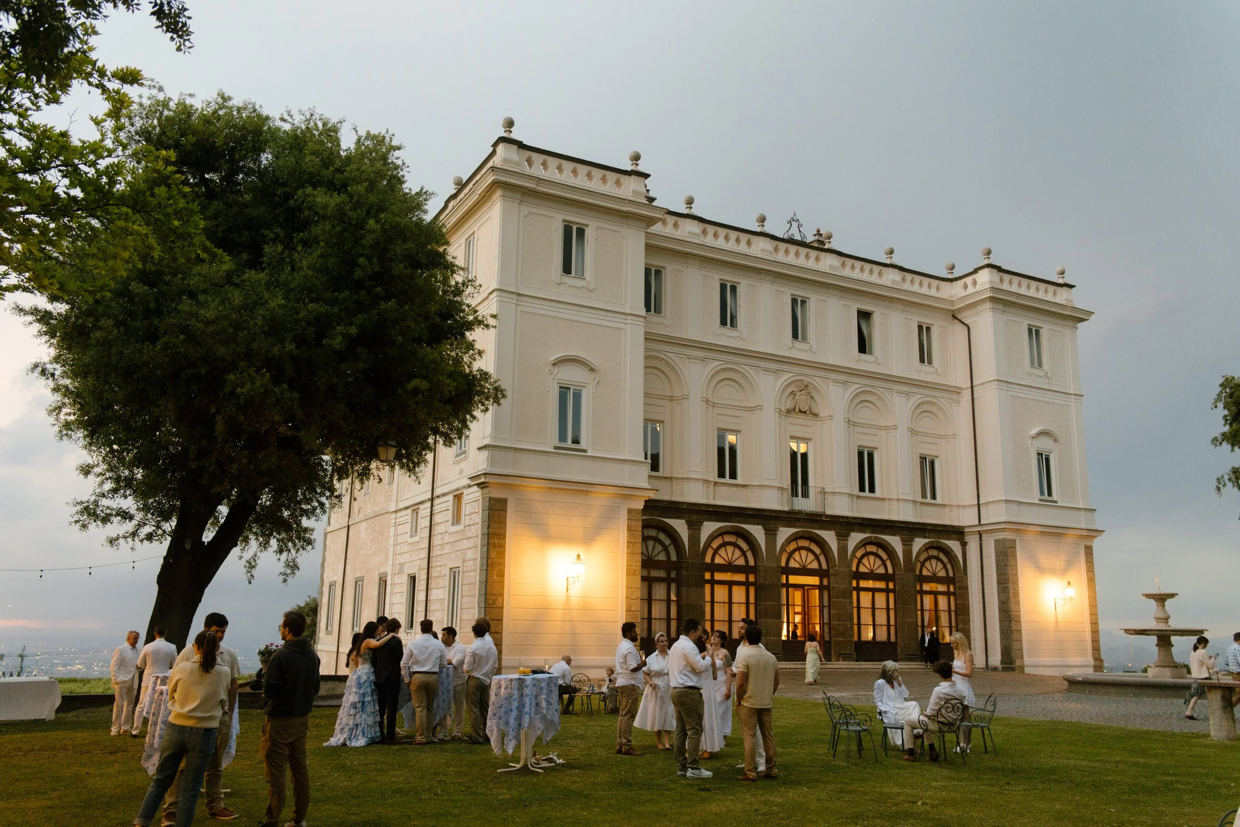 Guests gather on the lawn of a stunning Italian villa as twilight settles in, mingling beneath string lights and ancient trees. Effortless elegance at a wedding in Italy.