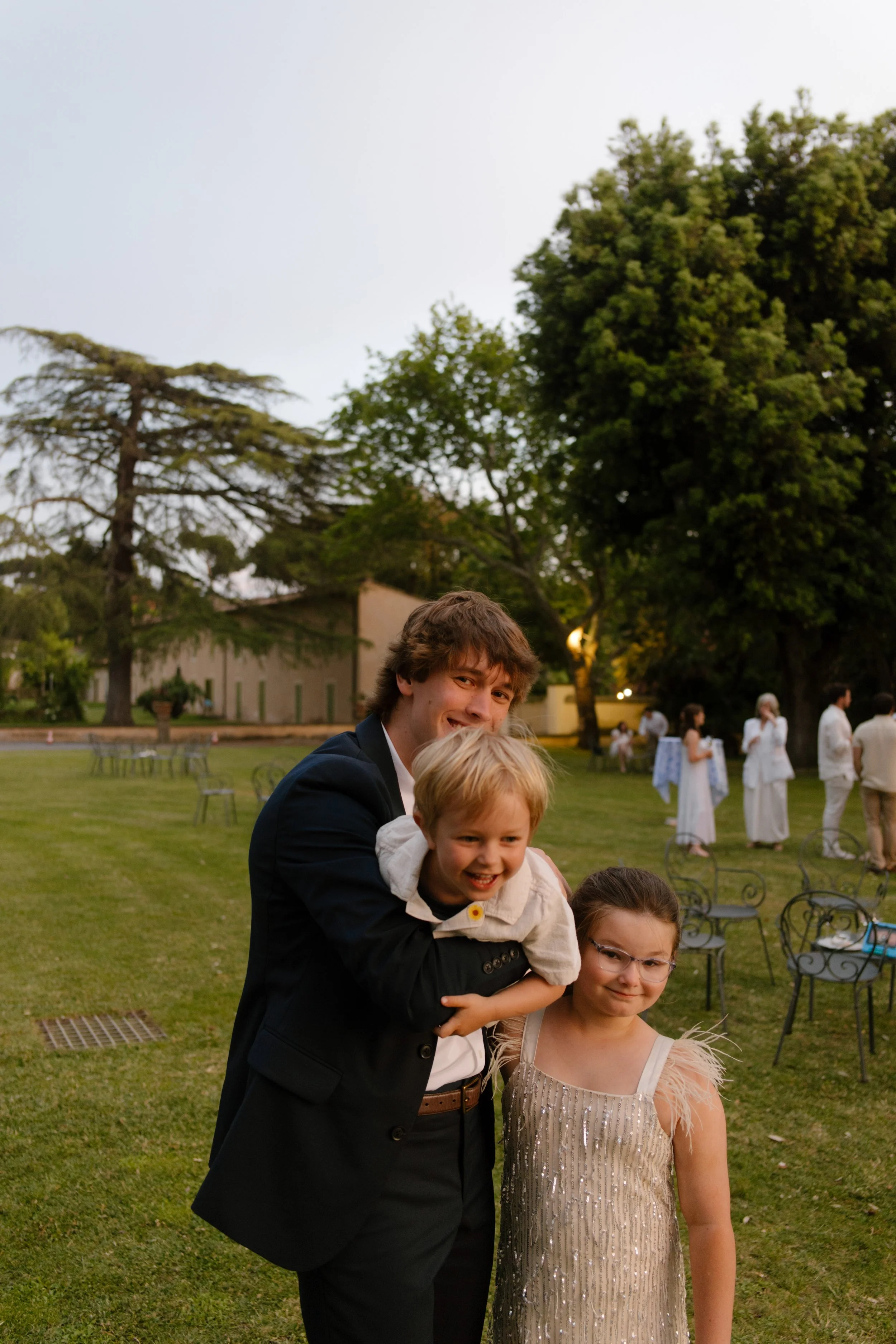 The groom crouches down to hug two giggling kids, dressed in their best with grass-stained joy in their eyes. Real life, real love, right in the middle of the party.