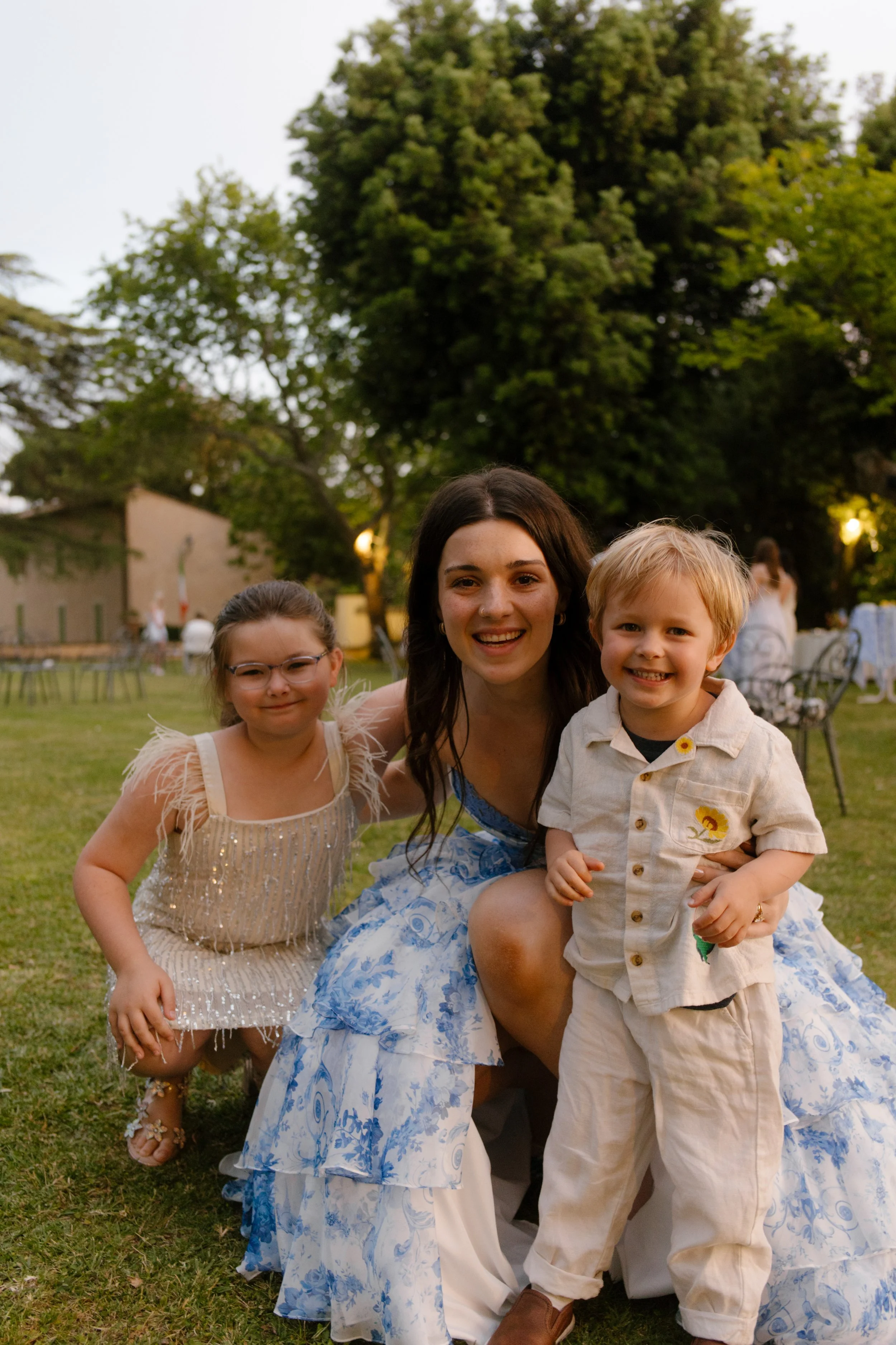 The bride kneels on the grass with two flower kids wrapped around her, all smiles and sparkle. Post-ceremony cuddles captured during a whimsical wedding in Italy.