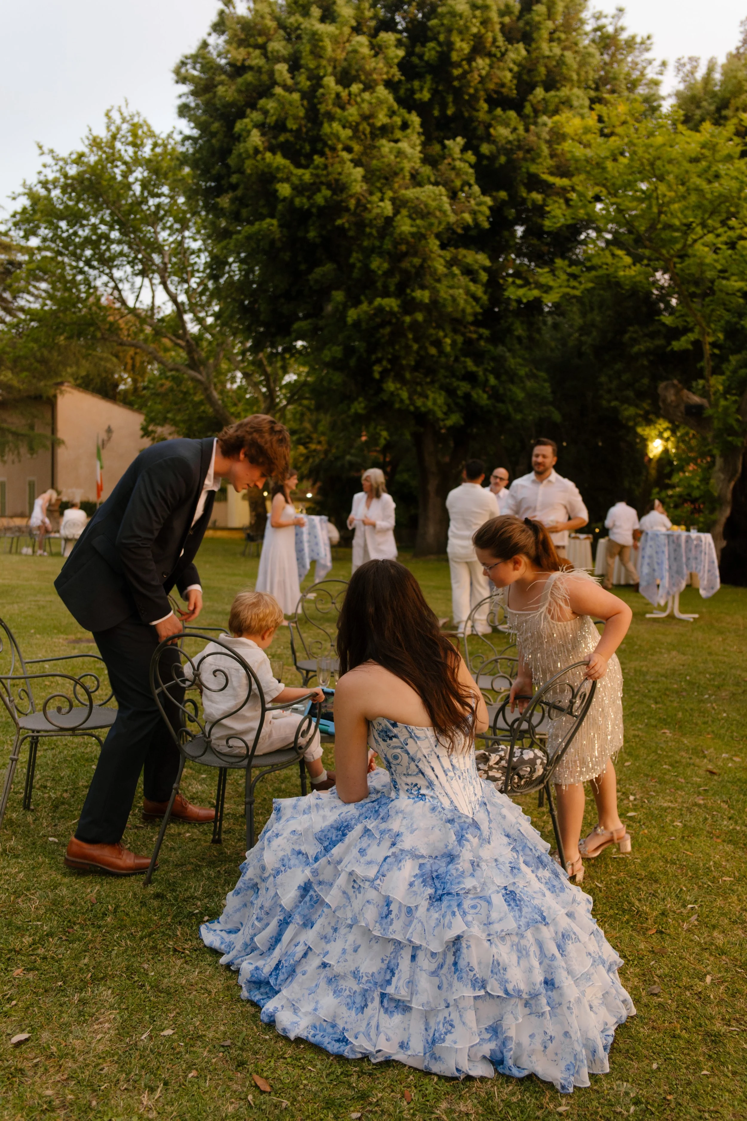 The couple interacts with children seated on vintage iron chairs during a reception on the lawn, white outfits and laughter all around. Intimate in-between moments that make the memory.